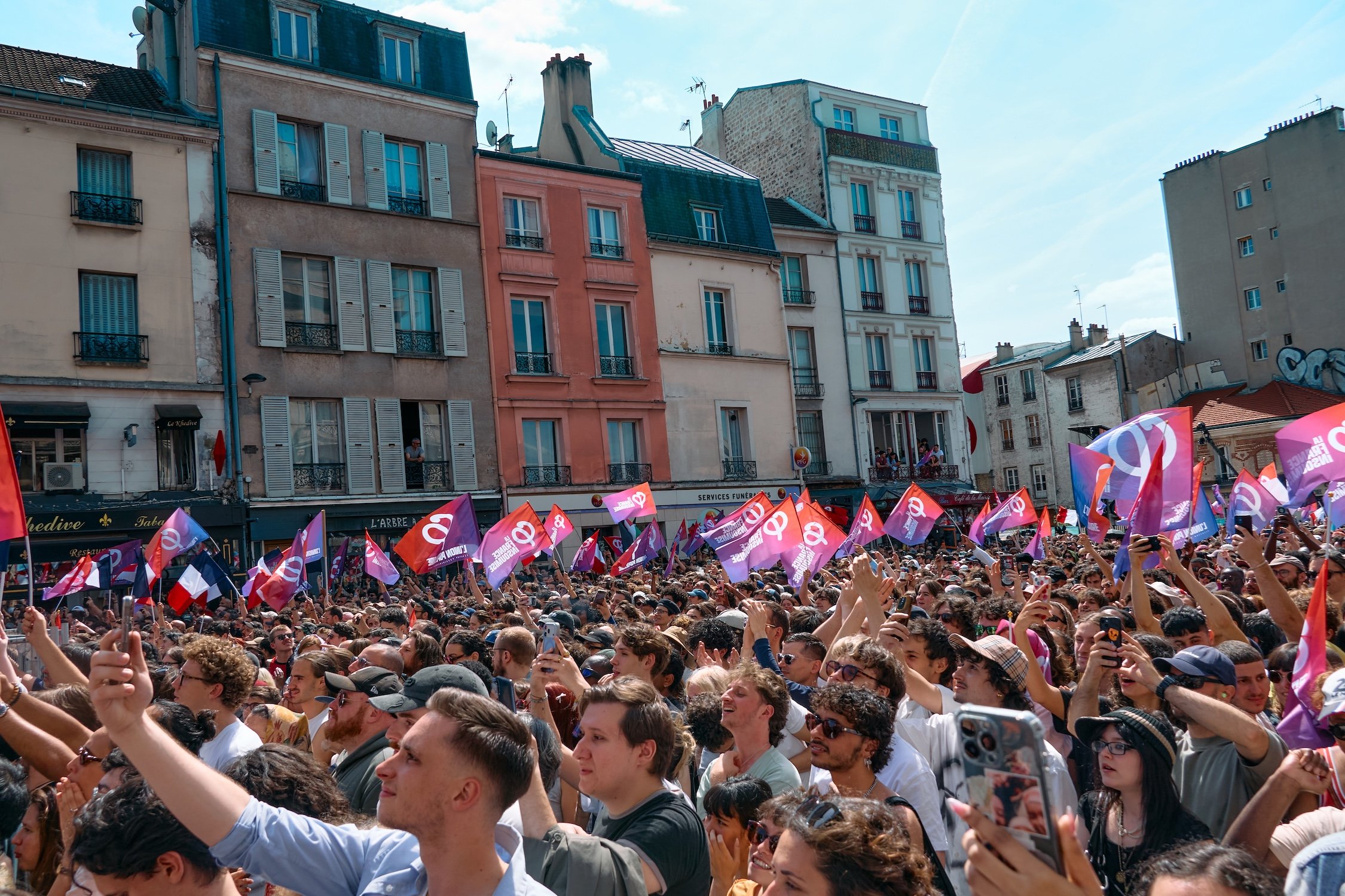 A large crowd of people gathered outdoors during a sunny day, many holding pink and blue flags with white symbols, in front of old European style buildings with multiple stories and balconies.