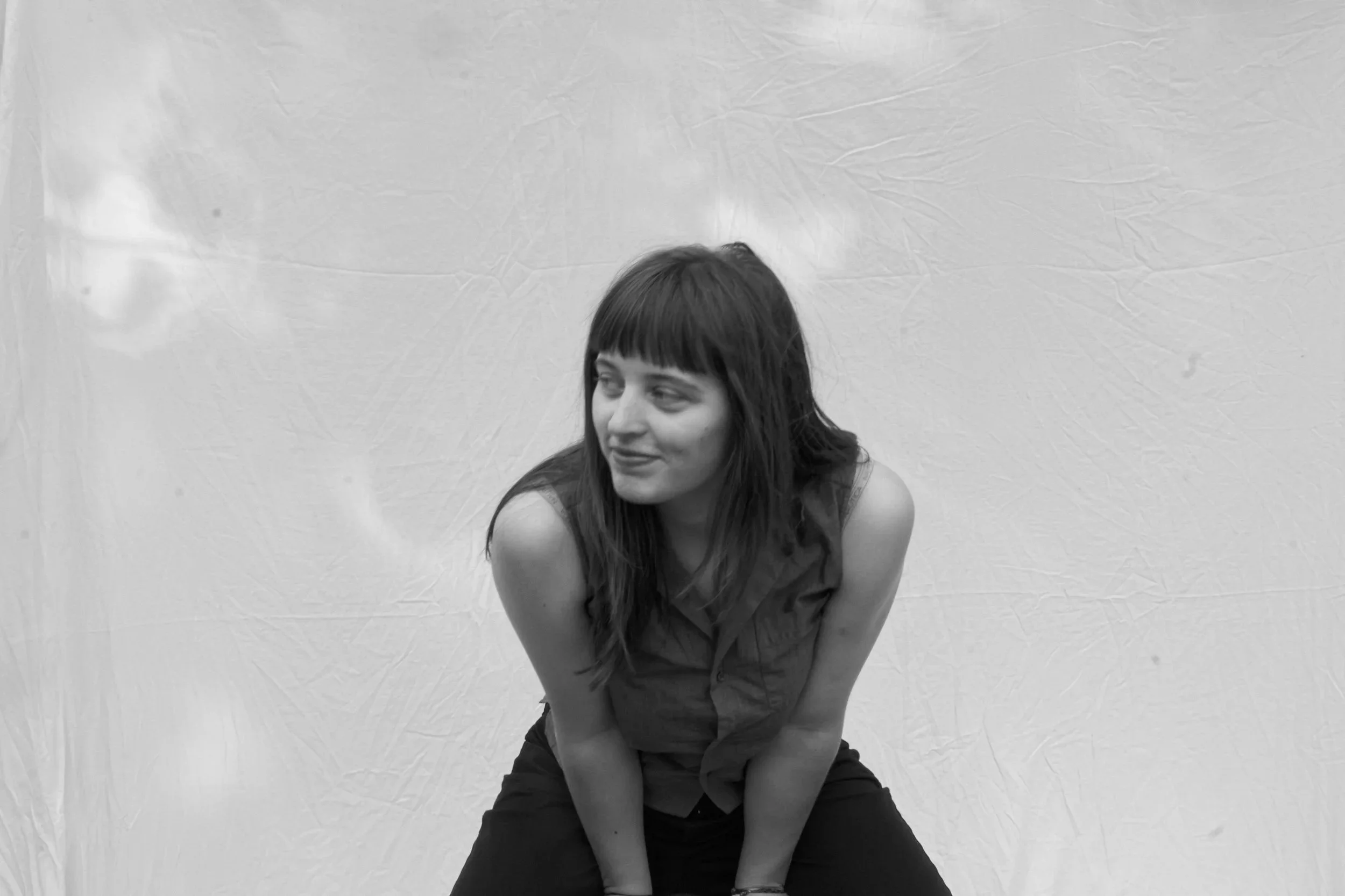 A woman with dark hair and bangs, wearing a sleeveless top, seated against a wrinkled cloth backdrop, looking to her right with a subtle smile.