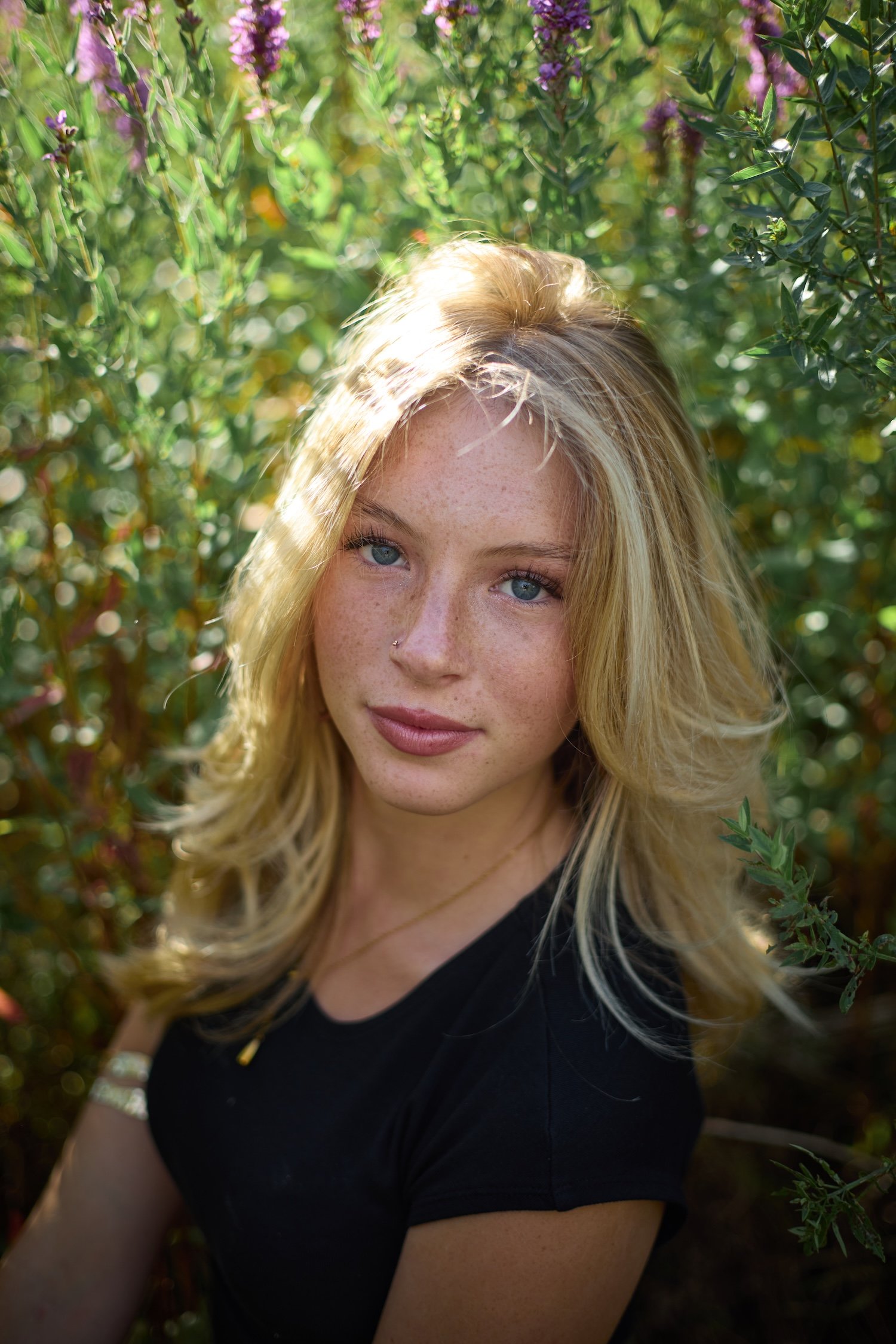 Close-up of a young woman with long blonde hair and freckles, wearing a black shirt, standing outdoors among green bushes with purple flowers, with sunlight highlighting her hair and face.