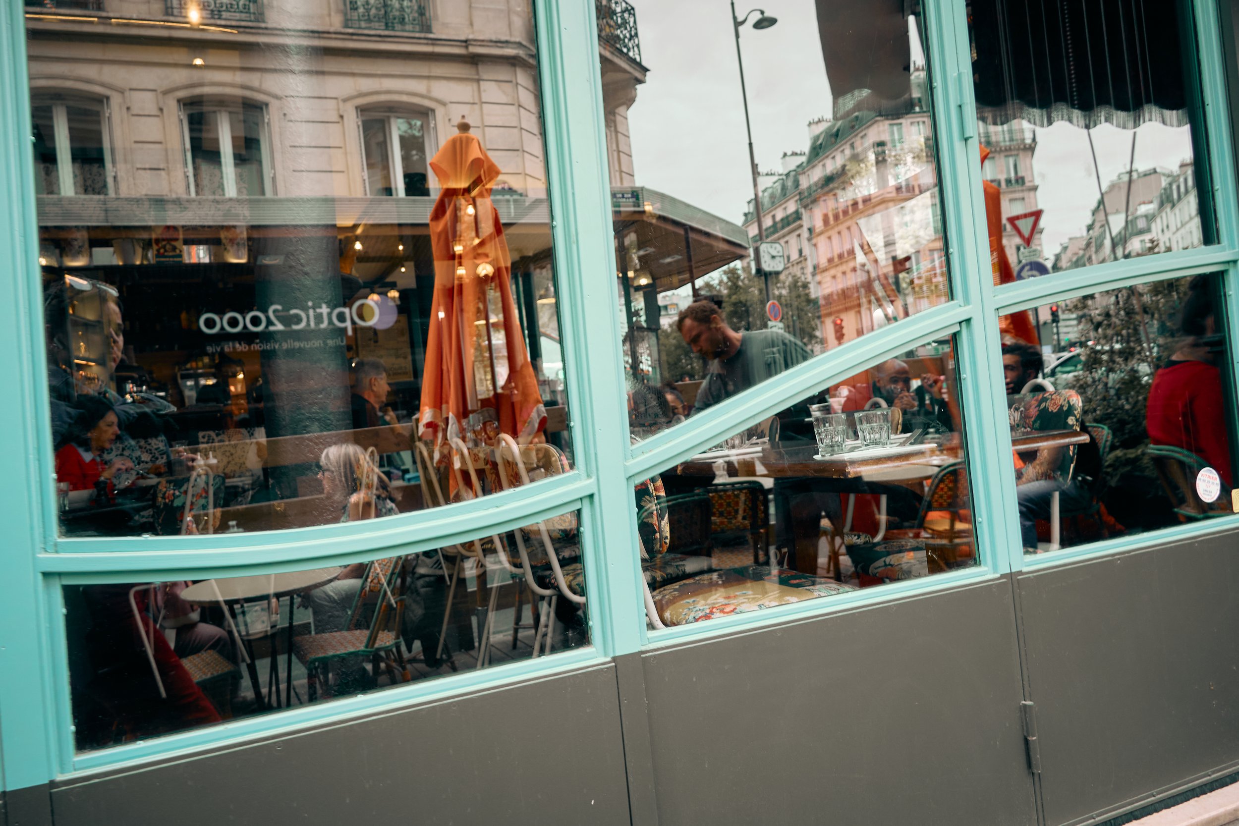 View through a large window of a busy café with people sitting and dining, orange umbrellas outside, and Parisian buildings in the background, reflecting city life.
