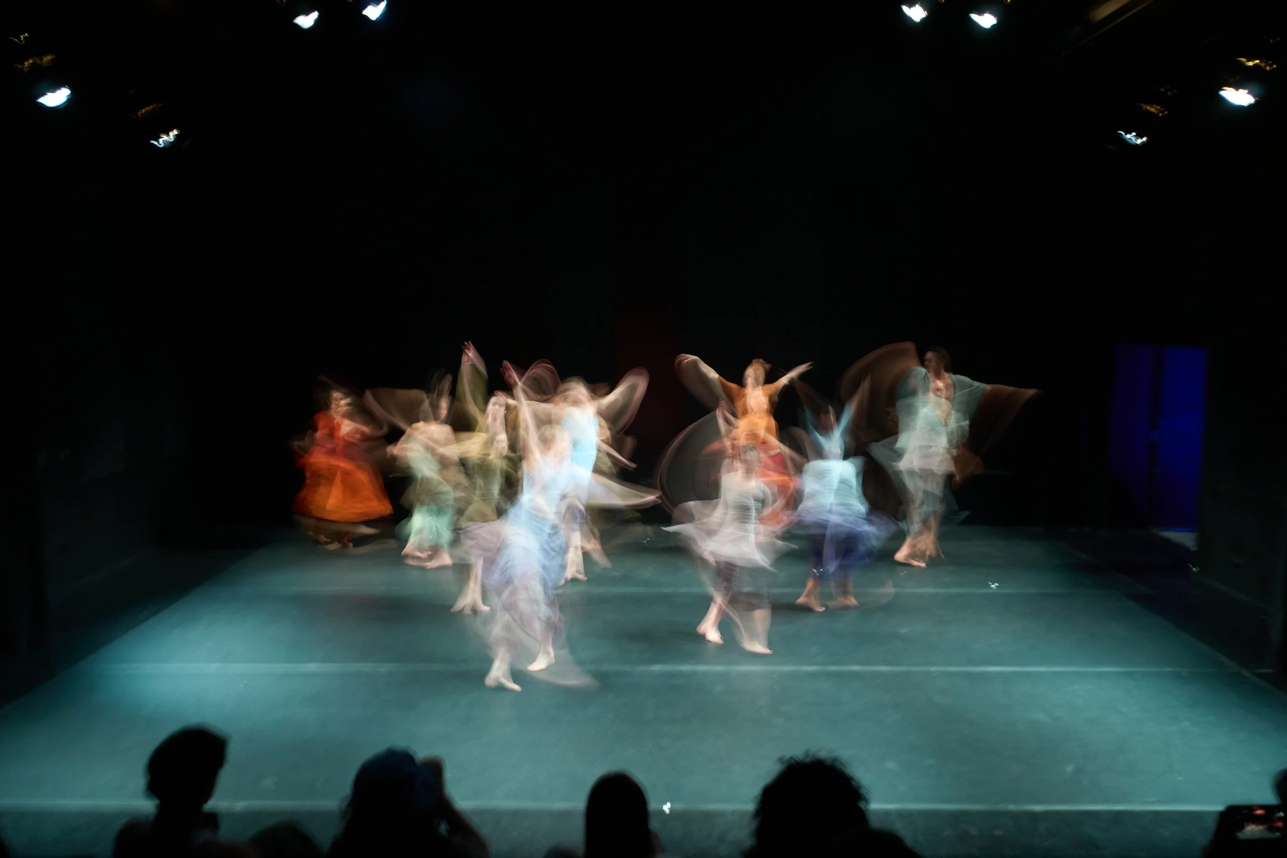 Blurred image of dancers performing on stage under bright spotlights, with audience visible at the front.