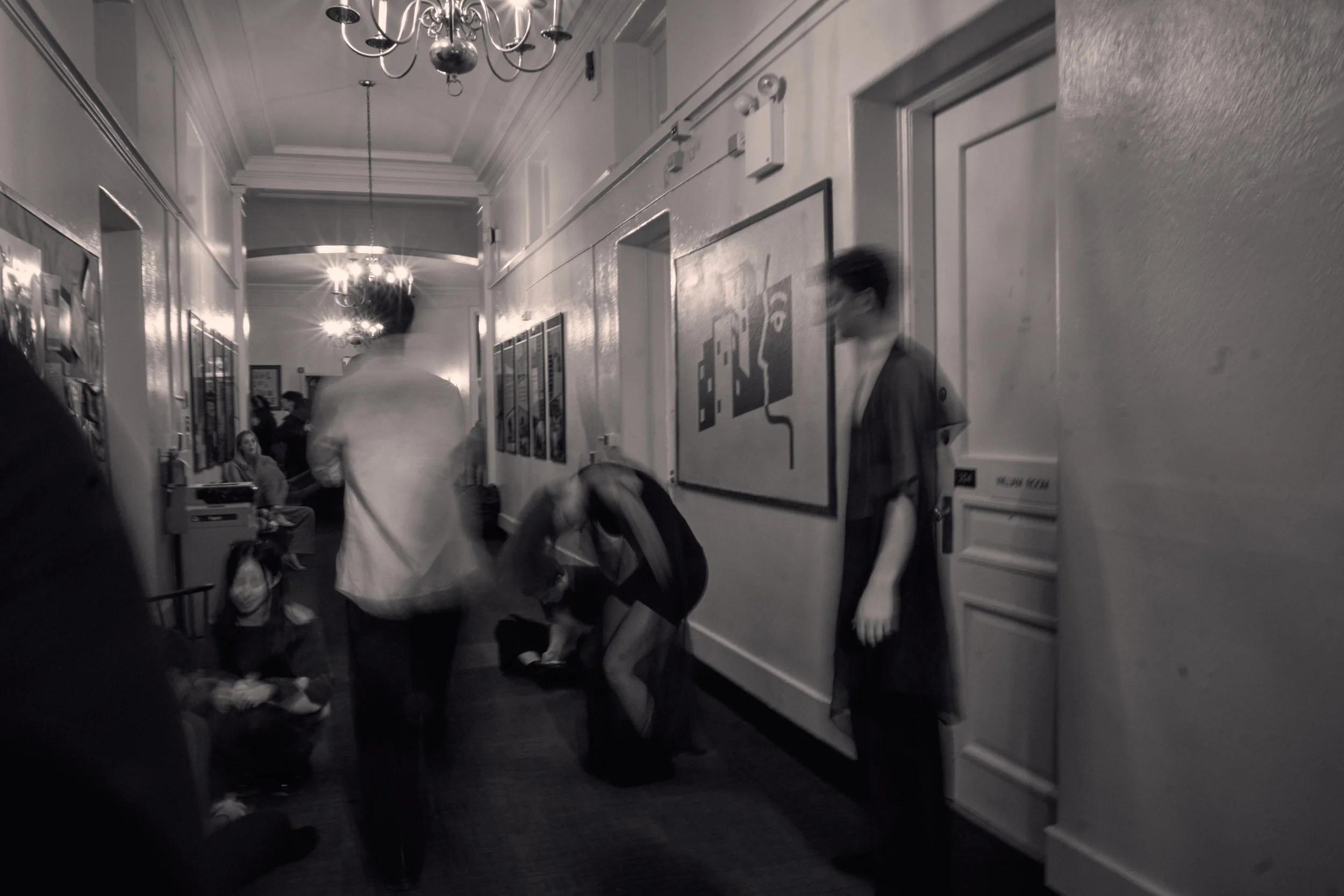 A hallway in a building with people walking. The hallway has ornate chandeliers hanging from the ceiling, and walls decorated with framed pictures and artwork. A woman wearing glasses and a jacket stands by the wall, and a man is seen walking with a 