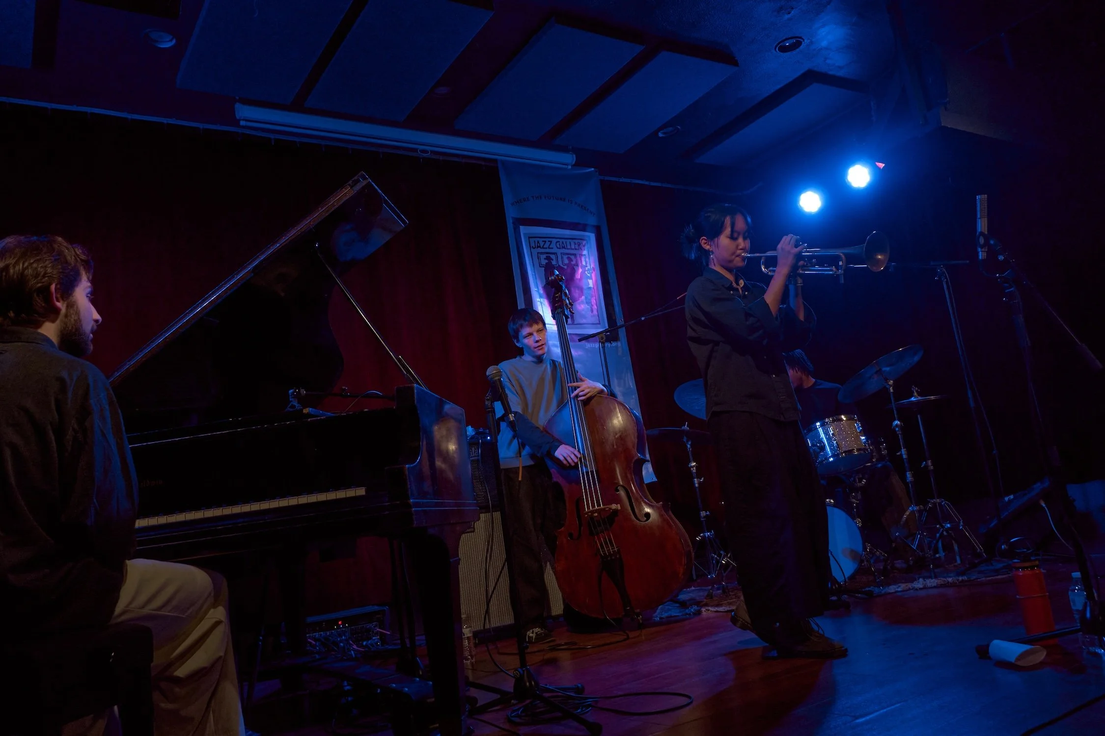 Jazz band performing on stage with piano, double bass, and drums, illuminated by blue stage lights.