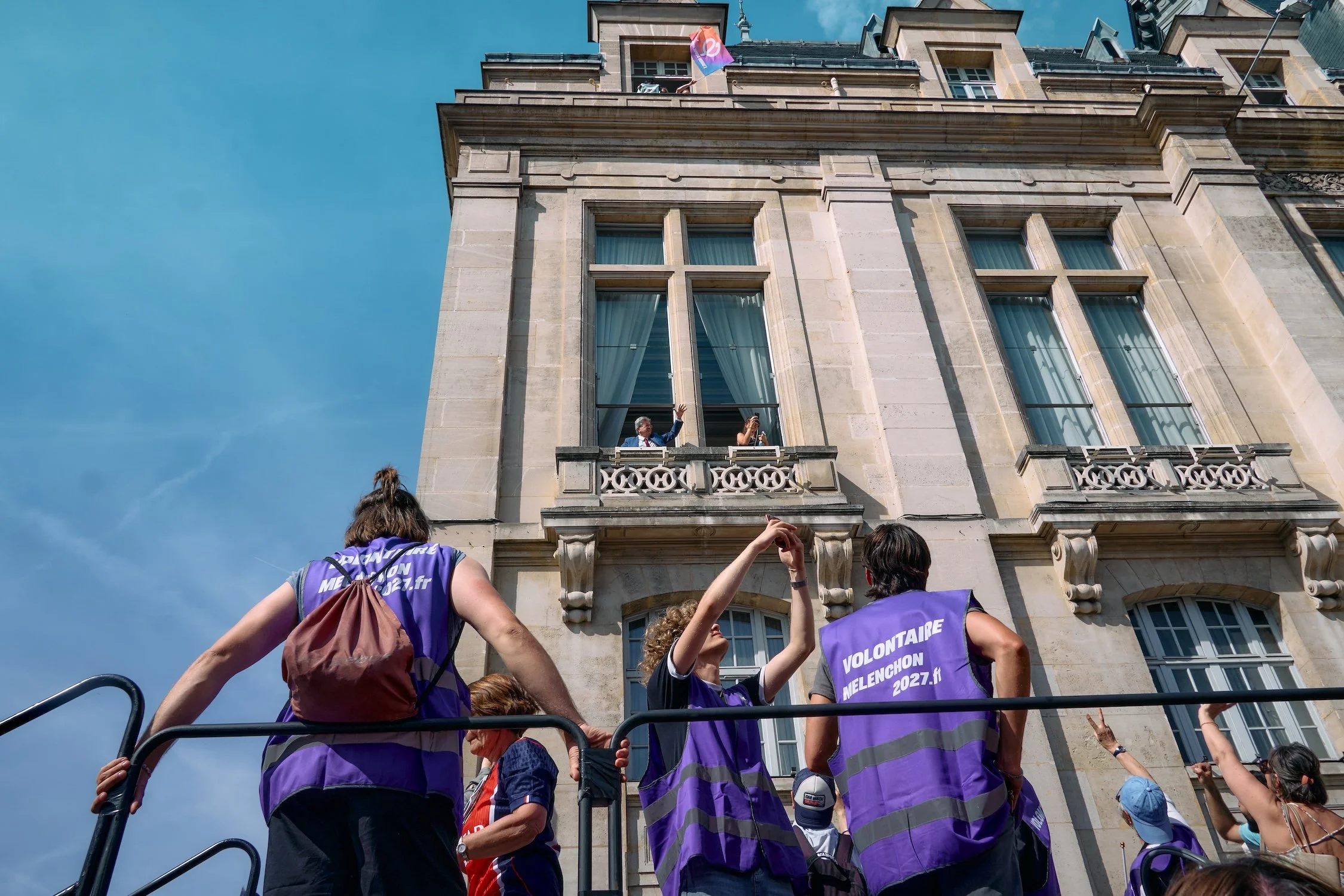 A group of people wearing purple vests with the words "VOLONTARI MELENCHON 2027.fr" on the back, raising their hands and looking up at a man in a suit and tie who is standing on a balcony of a large, historic building. The man on the balcony appears 