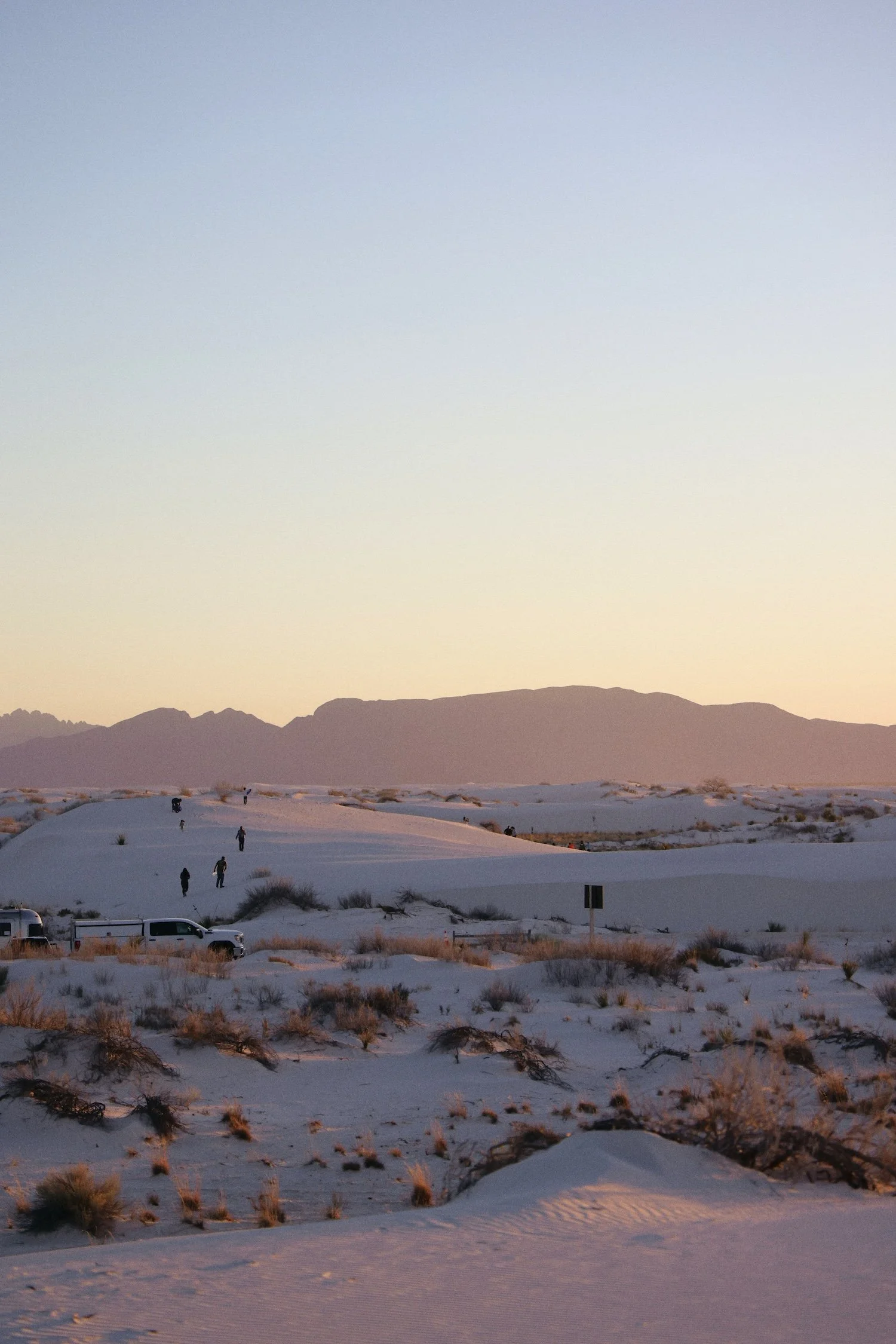 People hiking on sand dunes in a desert landscape with mountains in the background during sunset