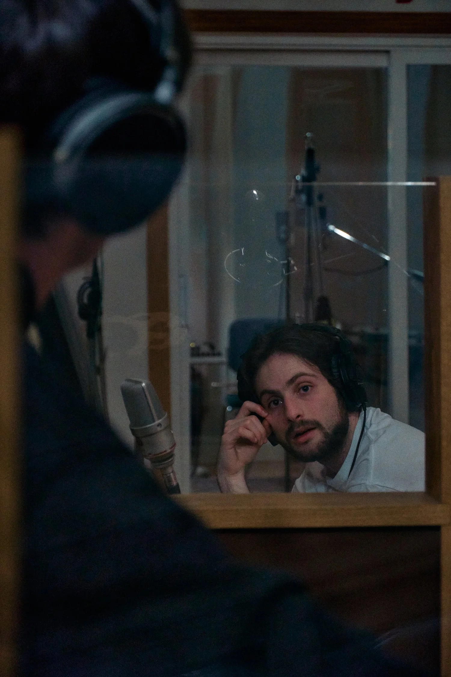 A man with dark hair and a beard wearing headphones is sitting in front of a microphone in a recording studio, while another person with dark hair and glasses is reflected in a glass window in the background.
