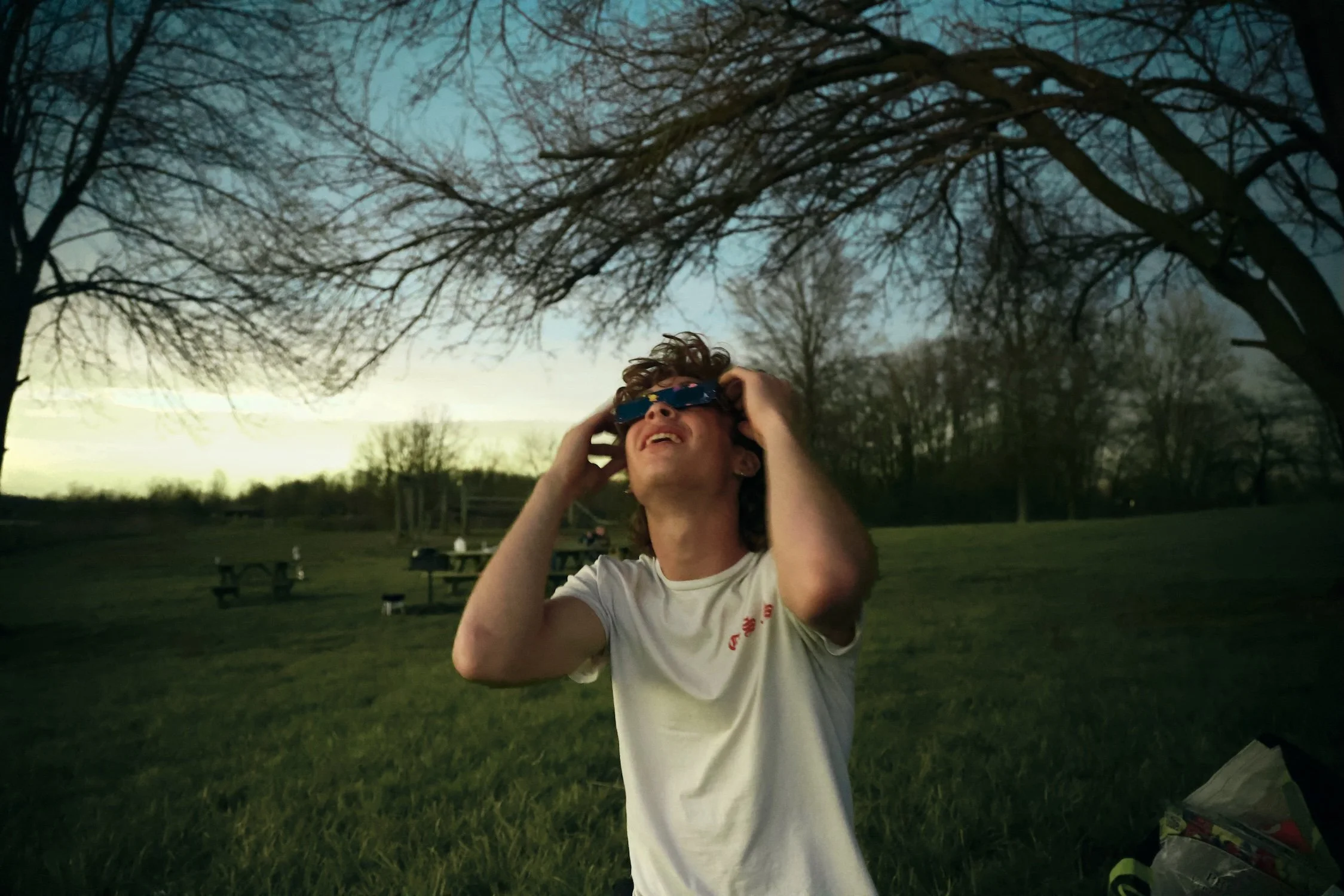 A person with curly hair and glasses is outdoors, wearing a white t-shirt, and adjusting their glasses while smiling. There are trees and picnic tables in the background during dusk or early evening.