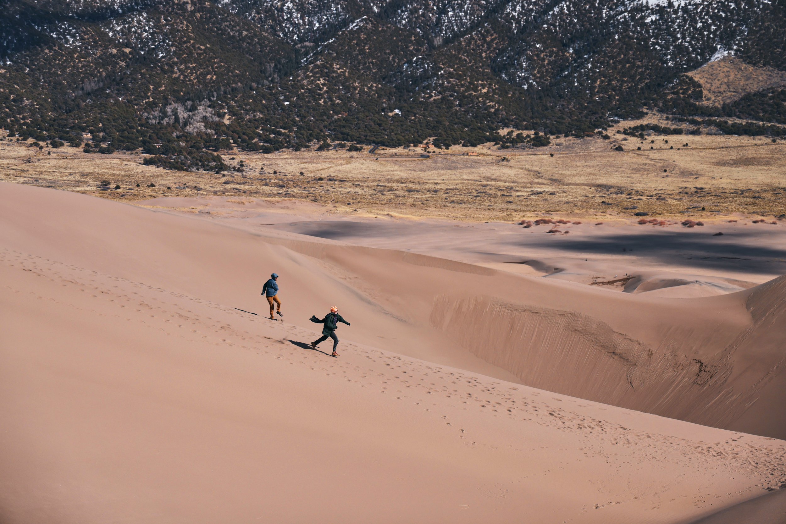 Three people walking on sand dunes in a desert landscape, with mountain hills in the background.