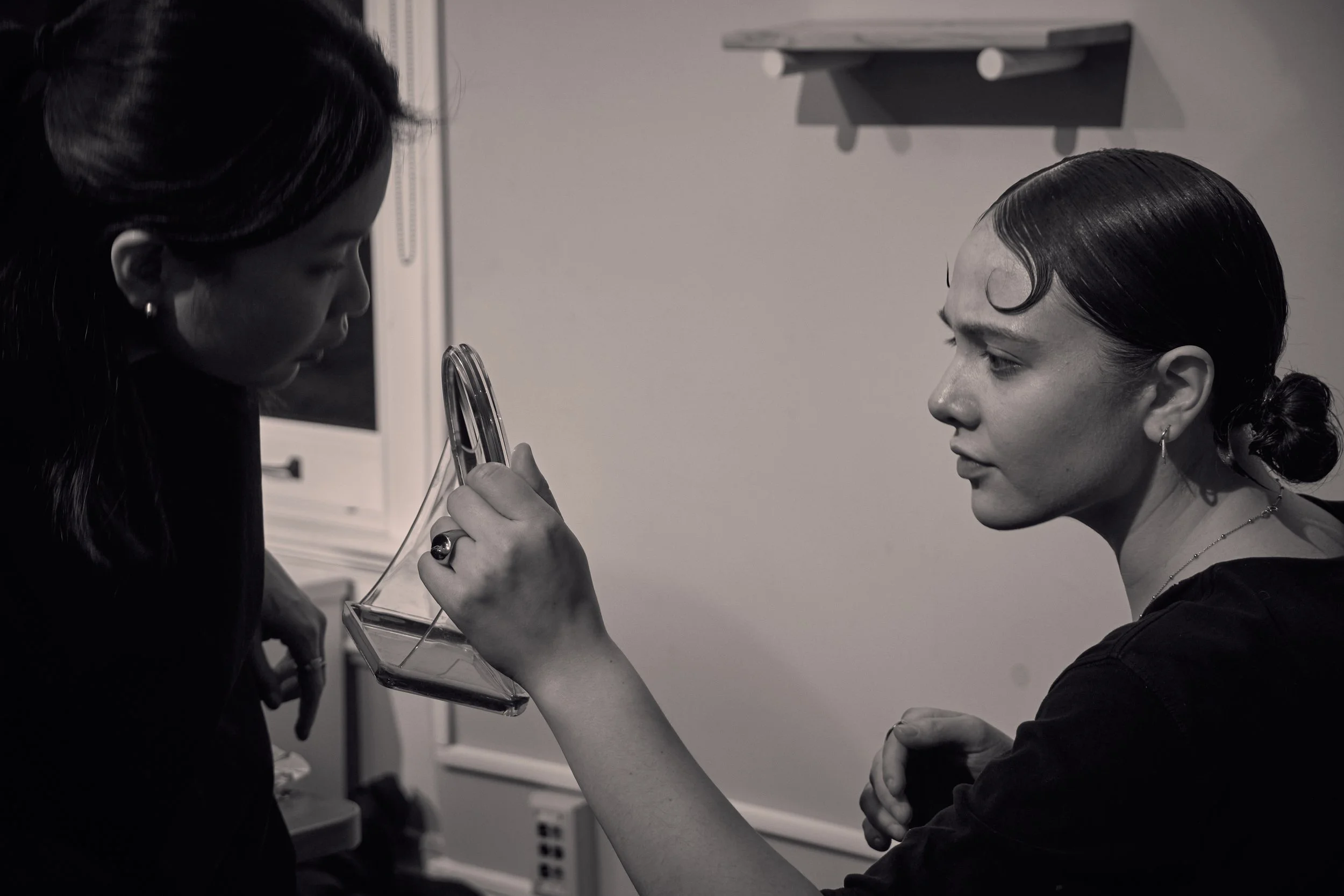 A woman with dark hair and earrings demonstrates a muscle flex to another woman with dark hair styled in an elegant bun, wearing a dark off-shoulder top, in a clinical or studio environment.