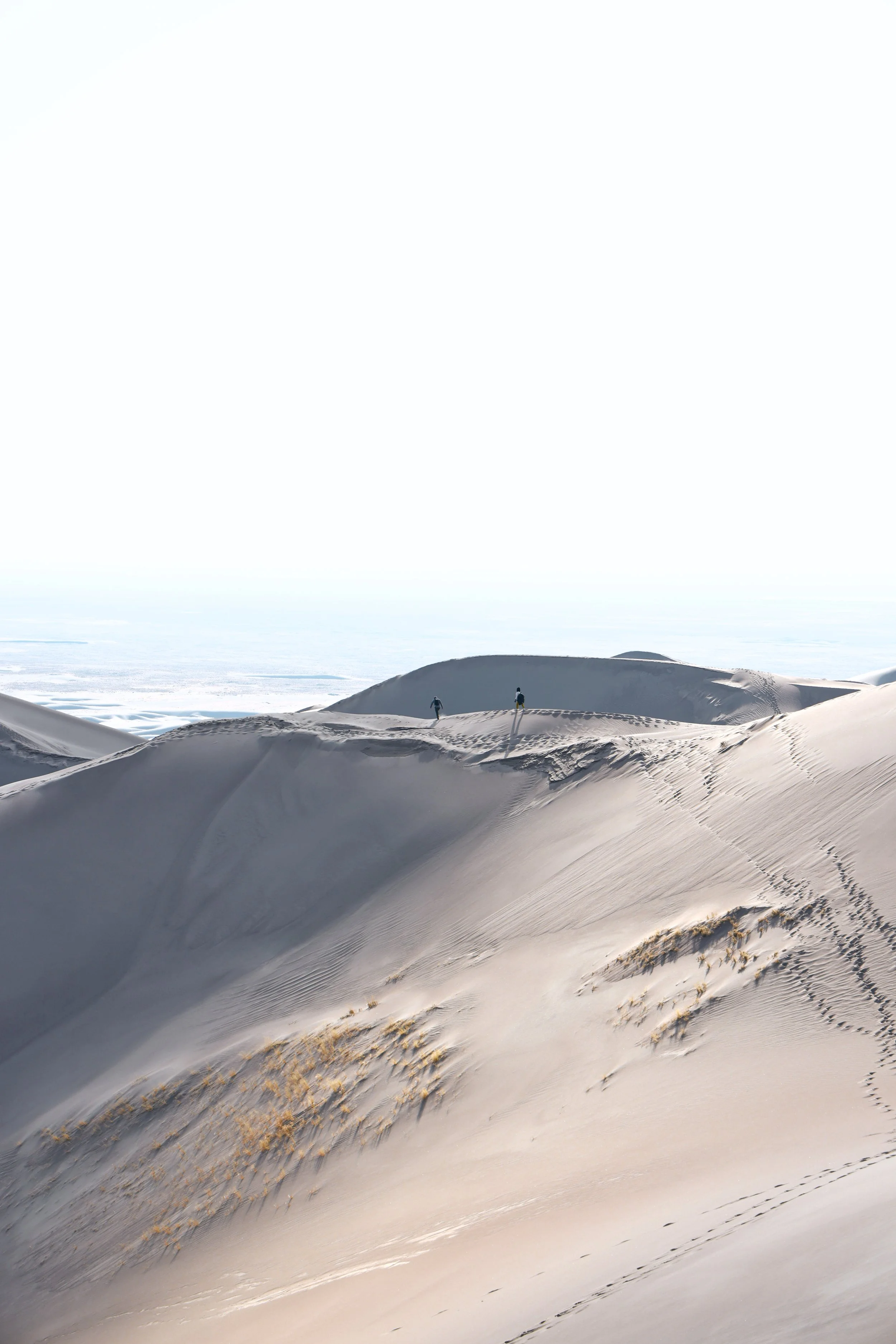 Snow-covered sand dunes under a bright, cloudy sky with two hikers walking across the dunes.