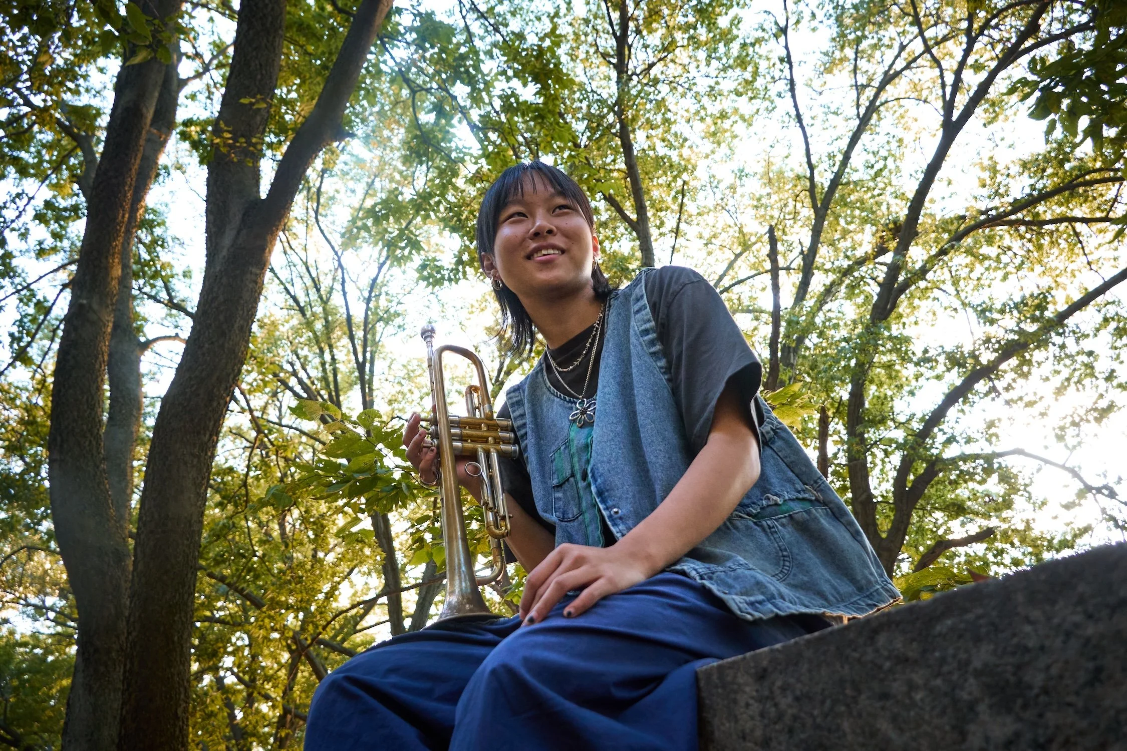 A young person sitting on a tree branch, holding a trumpet, surrounded by green leaves and trees during daylight.