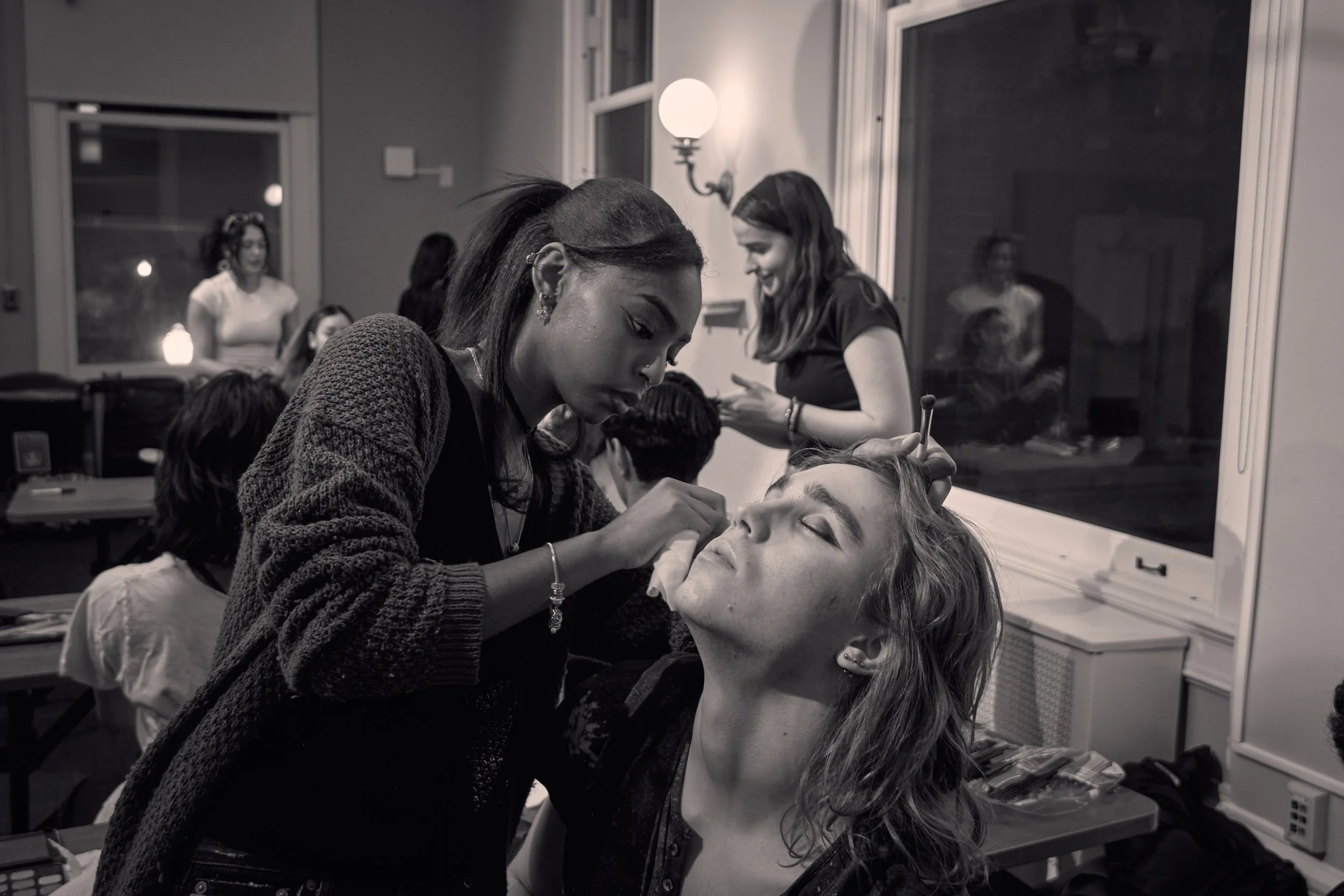 A makeup artist applying makeup to a woman's face in a room with other people, mirrors, and bright lighting.