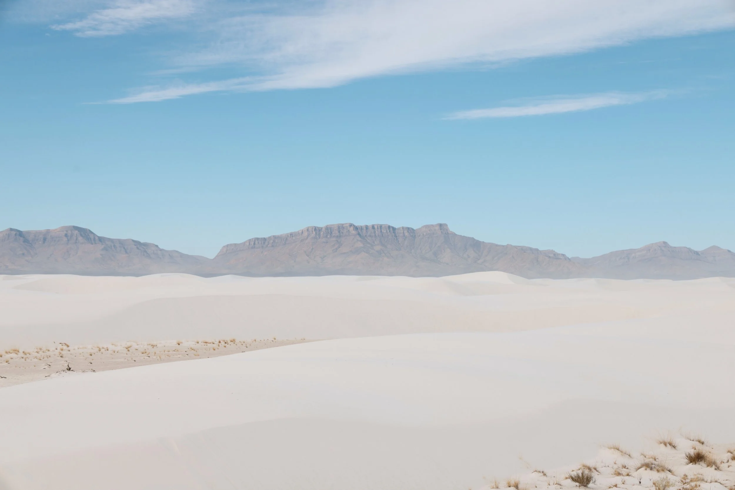 A vast desert landscape with white sand dunes, distant mountain ranges, and a clear blue sky with wispy clouds.