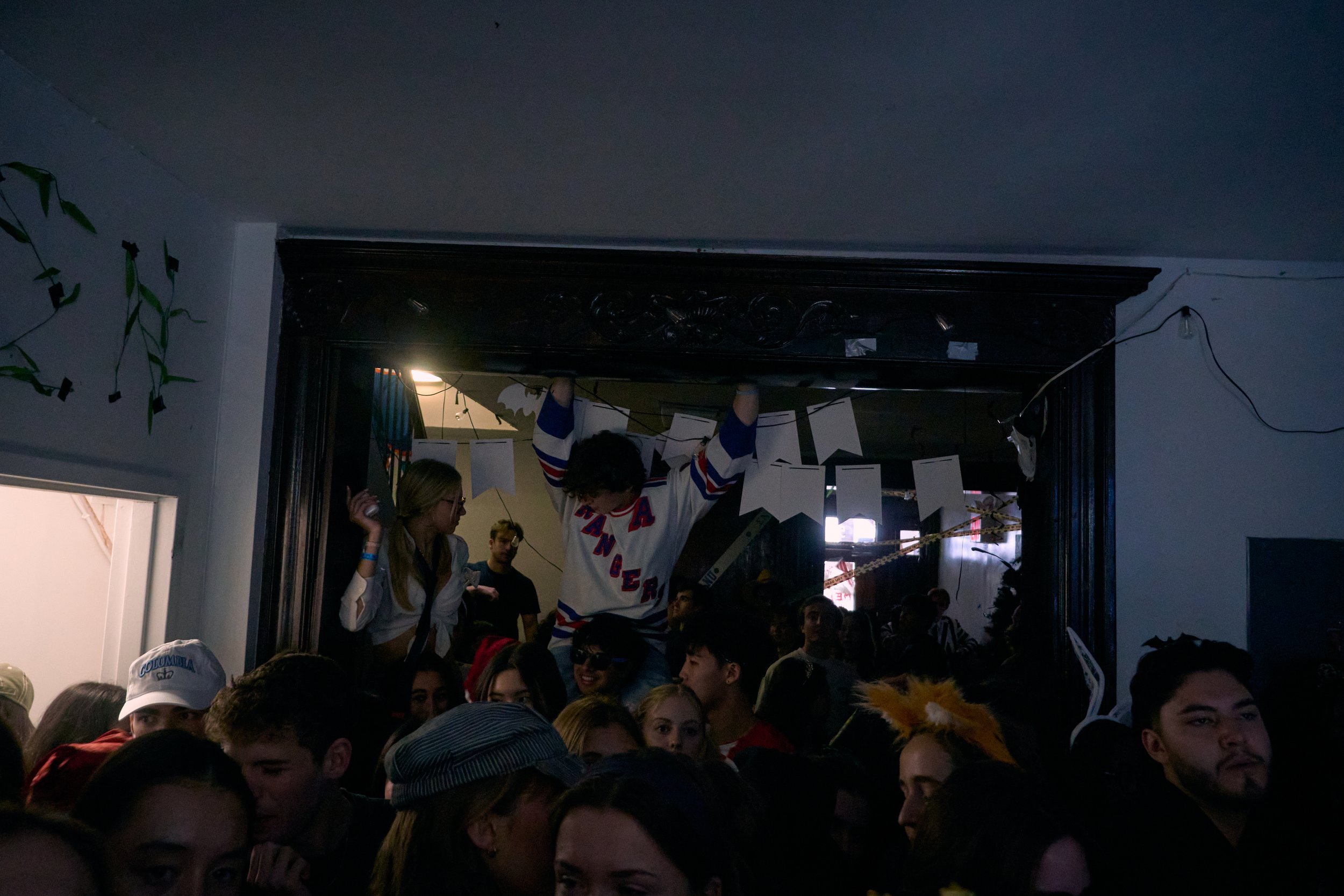 A large crowded room with many people, some wearing costumes and hats, appear to be at a party or gathering. Some individuals are on a raised platform or stage, engaging with the crowd. There are decorations hanging from the ceiling. The room is diml