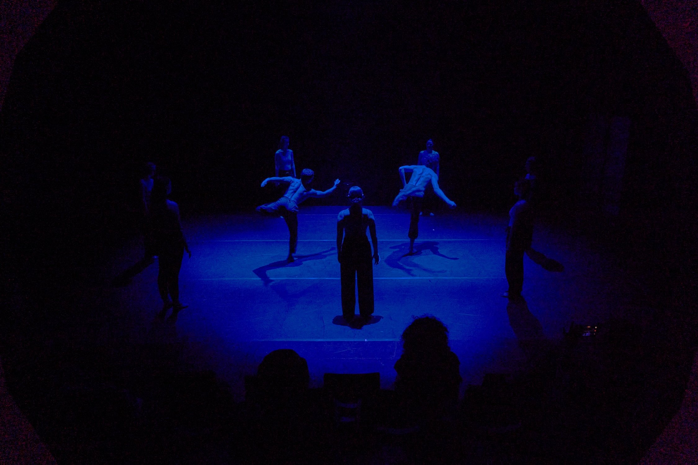 A group of dancers performing on a dark stage illuminated by blue and purple lights, with some dancers bending, standing, or sitting.