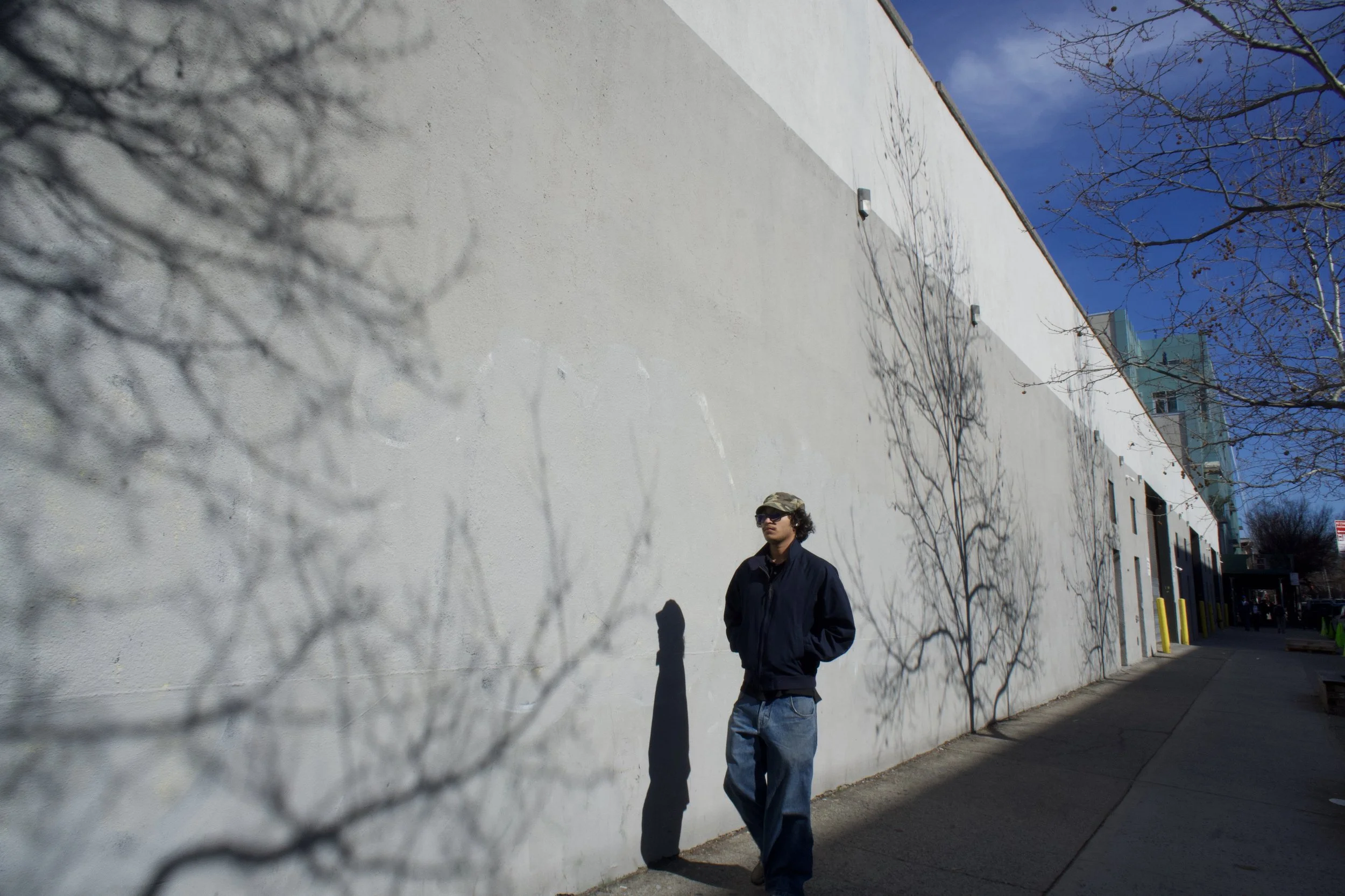 A young man with glasses and a cap standing on a sidewalk next to a white wall with shadows of trees cast on it, under a clear blue sky.