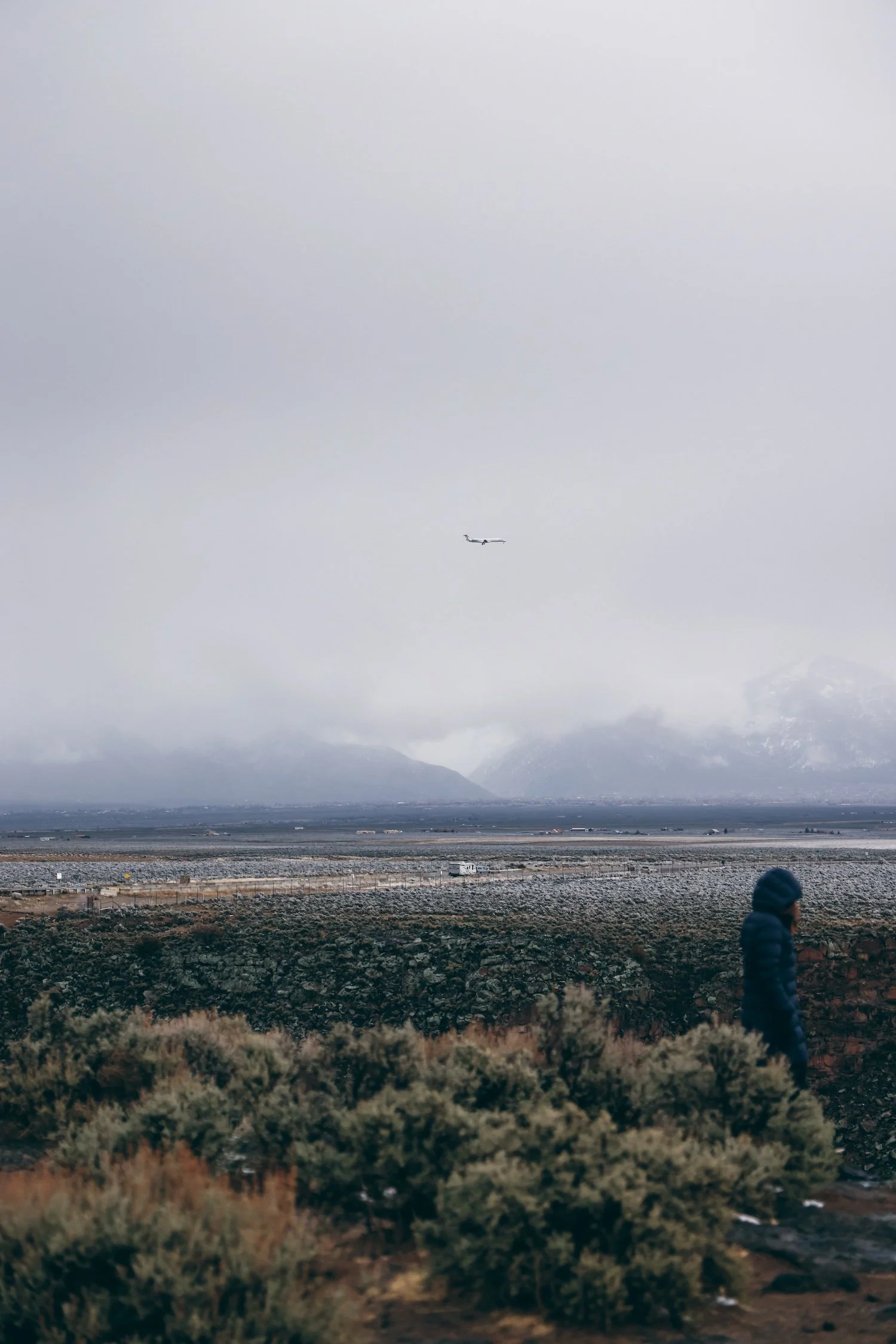 A landscape view with mountains in the background, a large body of water in the middle ground, and barren, rocky terrain with sparse bushes in the foreground. An airplane is flying in a cloudy sky, and a person in a dark jacket and hat is walking in 
