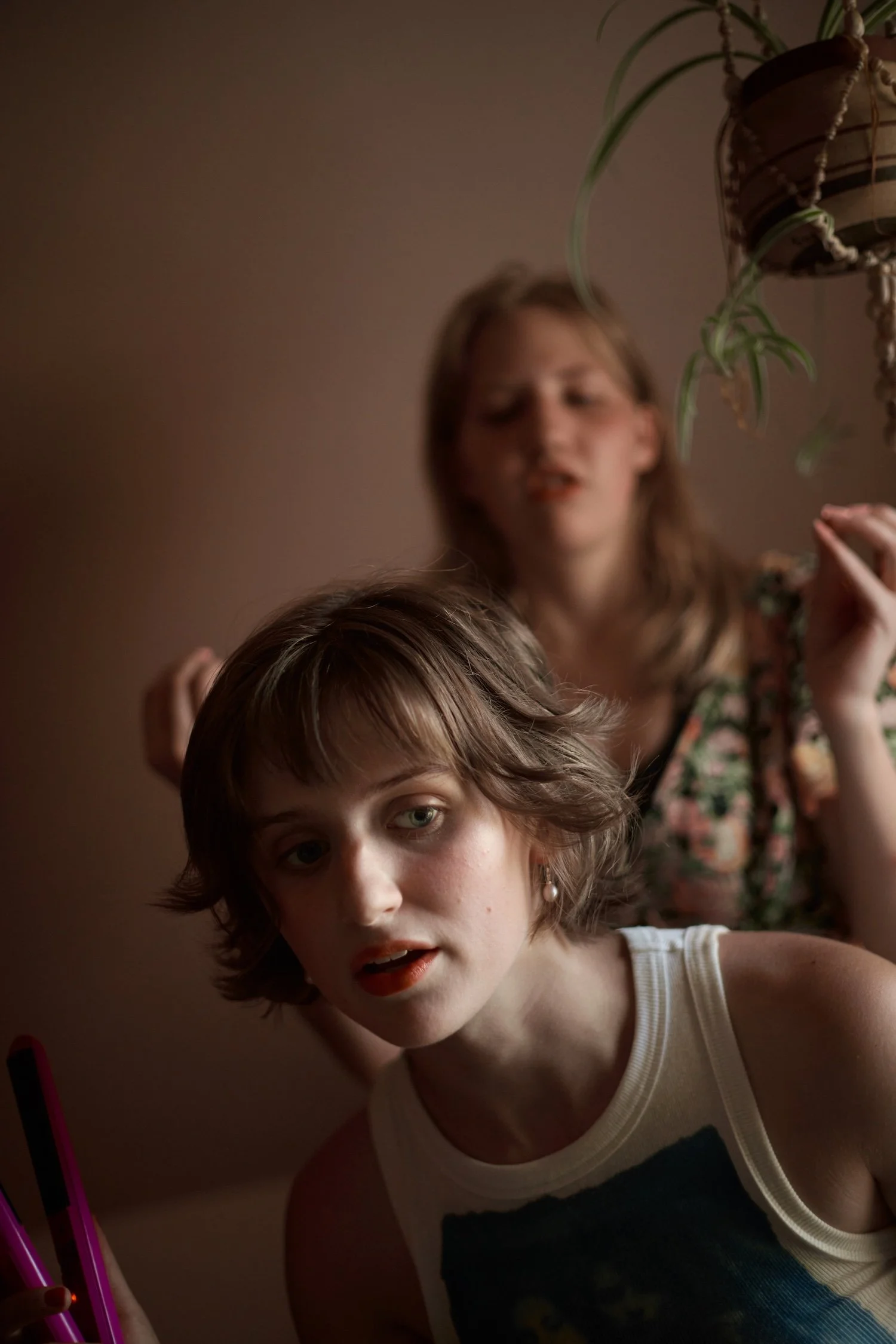 Young woman with short brown hair and green eyes wearing a white tank top with black and yellow graphic, posing in front of a beige wall, holding a pink glow stick.