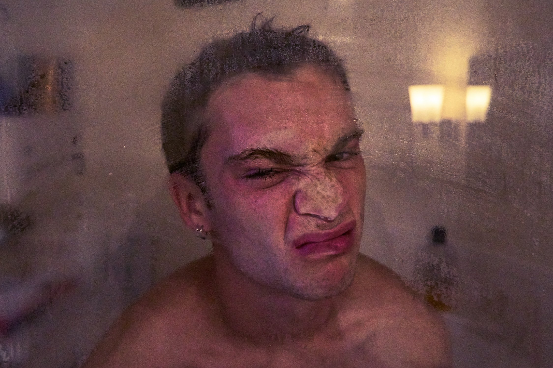 A woman with wet hair doing a playful face while taking a selfie behind a foggy or steamy glass