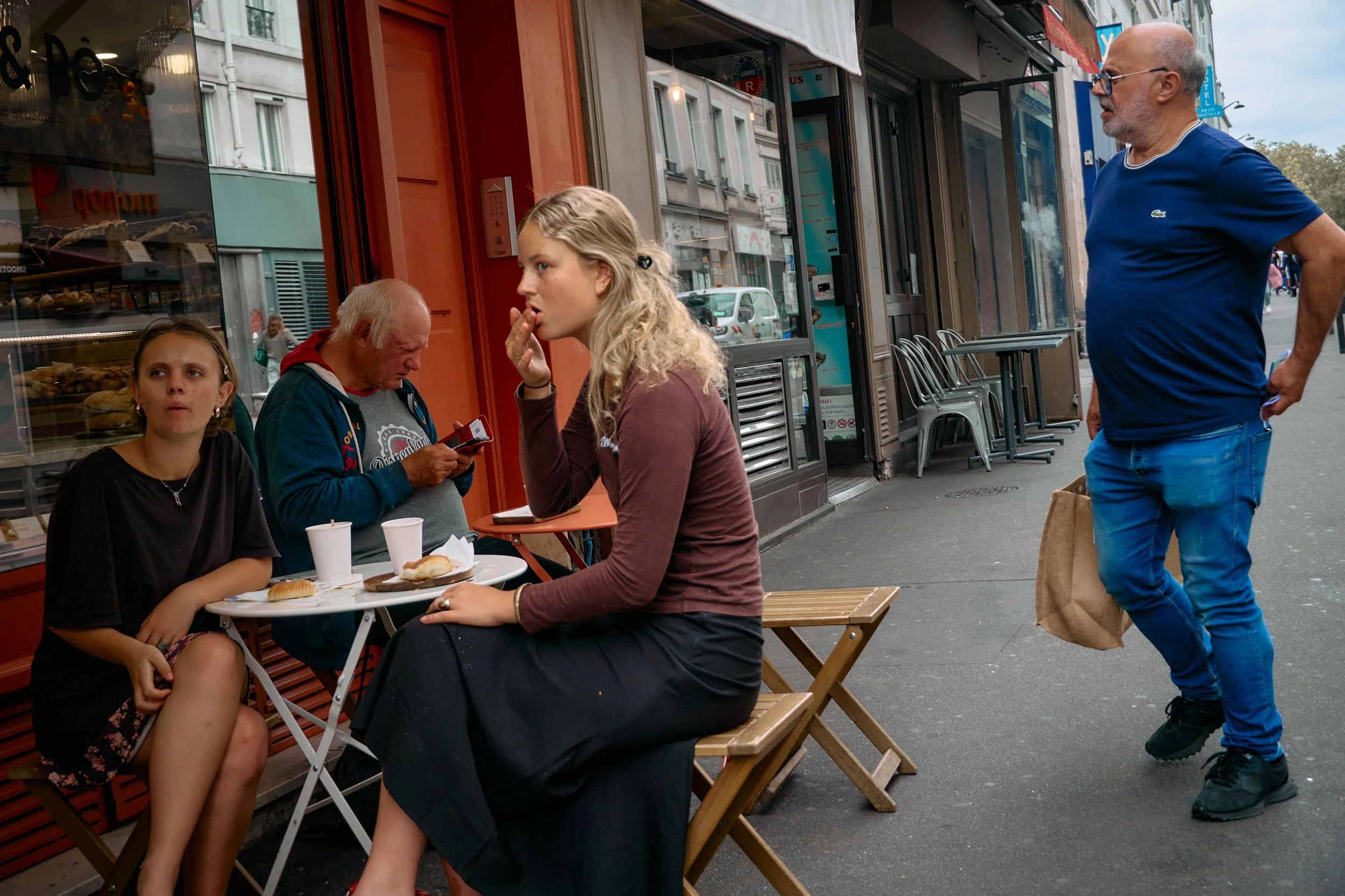 Four people are sitting and standing outside a bakery or cafe on a city sidewalk. Three women are seated at a small outdoor table with food, and one man is walking past them, carrying a brown paper bag.