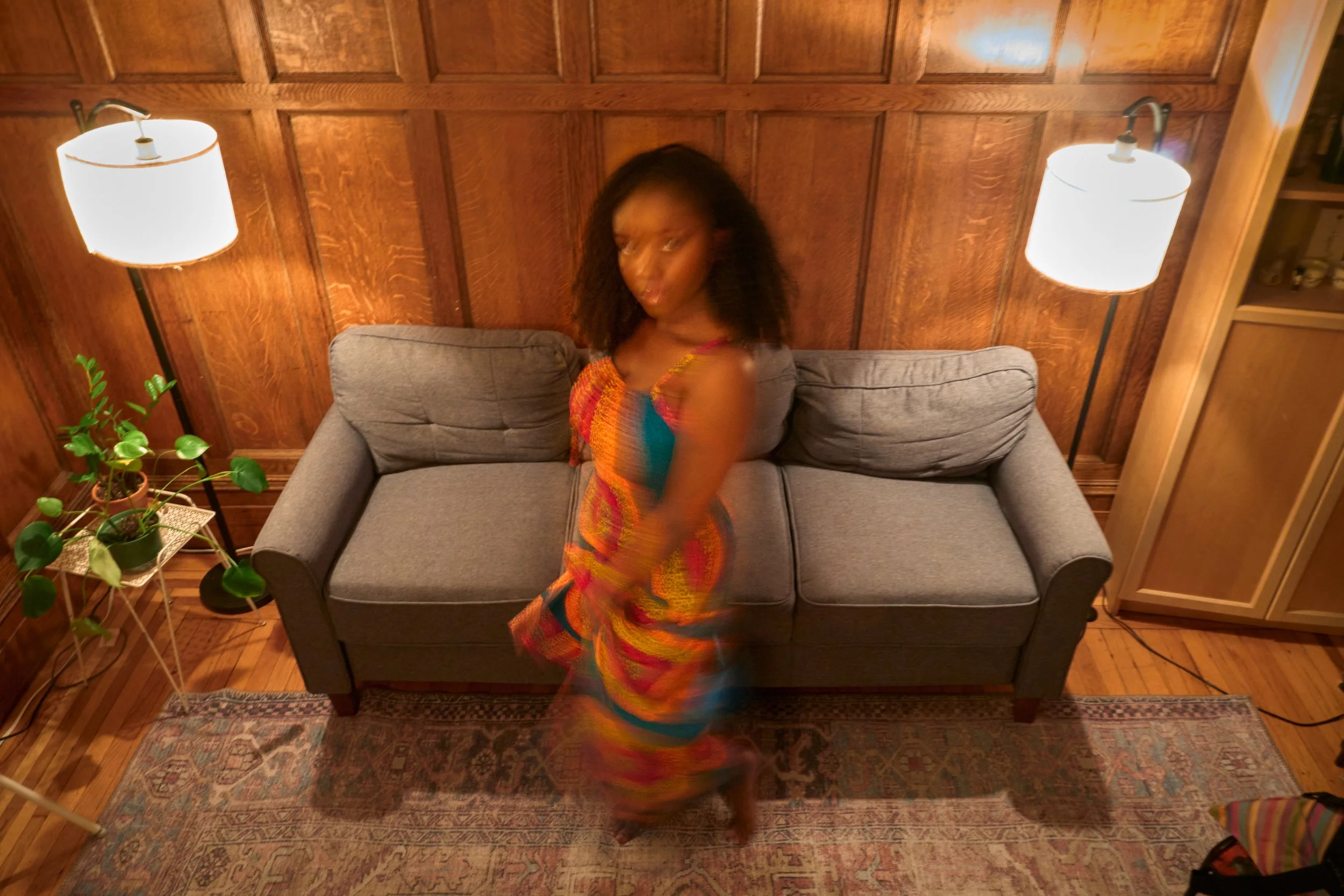 A woman in a colorful dress standing in front of a gray sofa in a room with wooden walls, two white wall-mounted lamps, and a plant on a small table to the left.