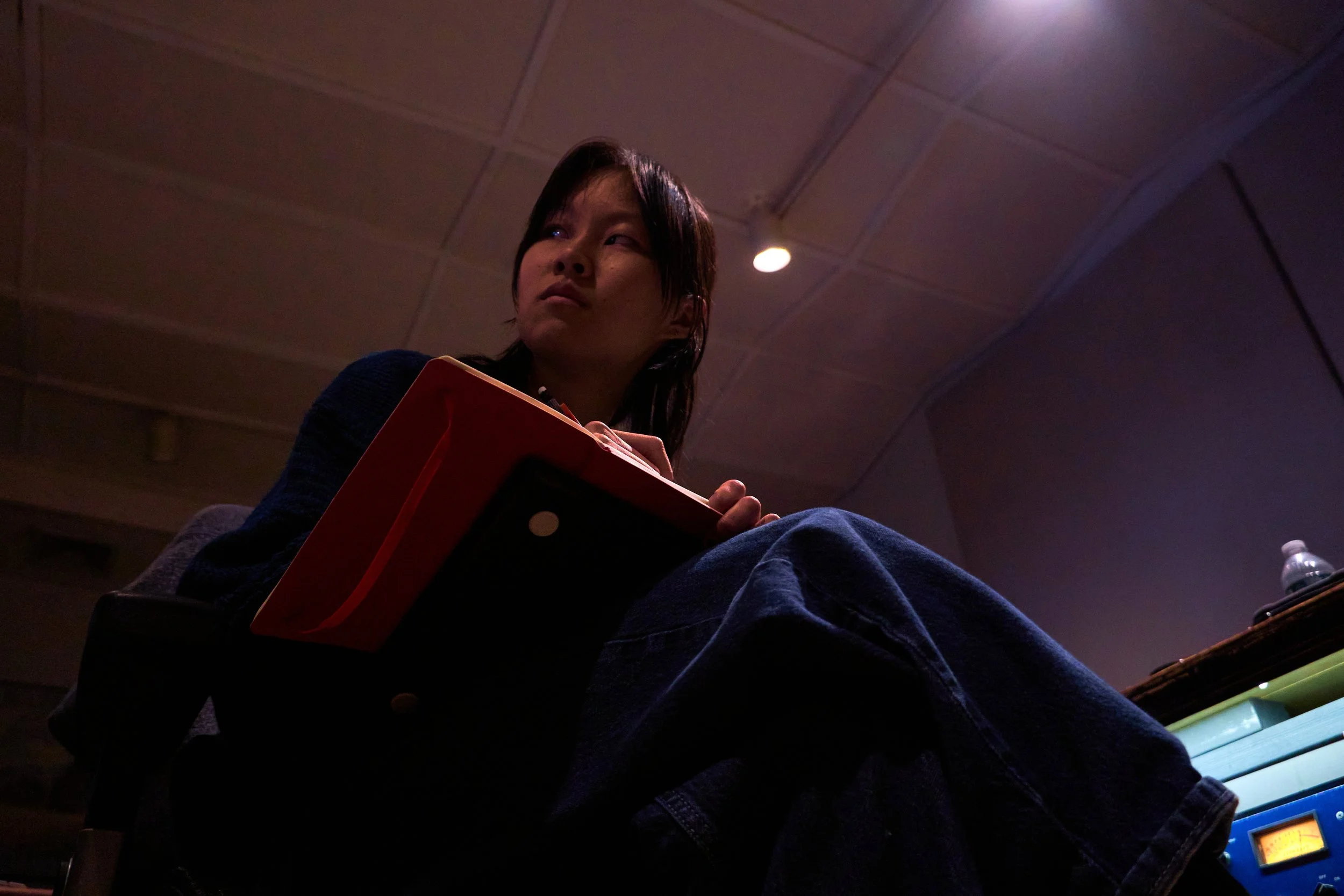 A young woman with dark hair sitting in a dimly lit room, holding a red notebook, with a water bottle and a calculator in front of her.