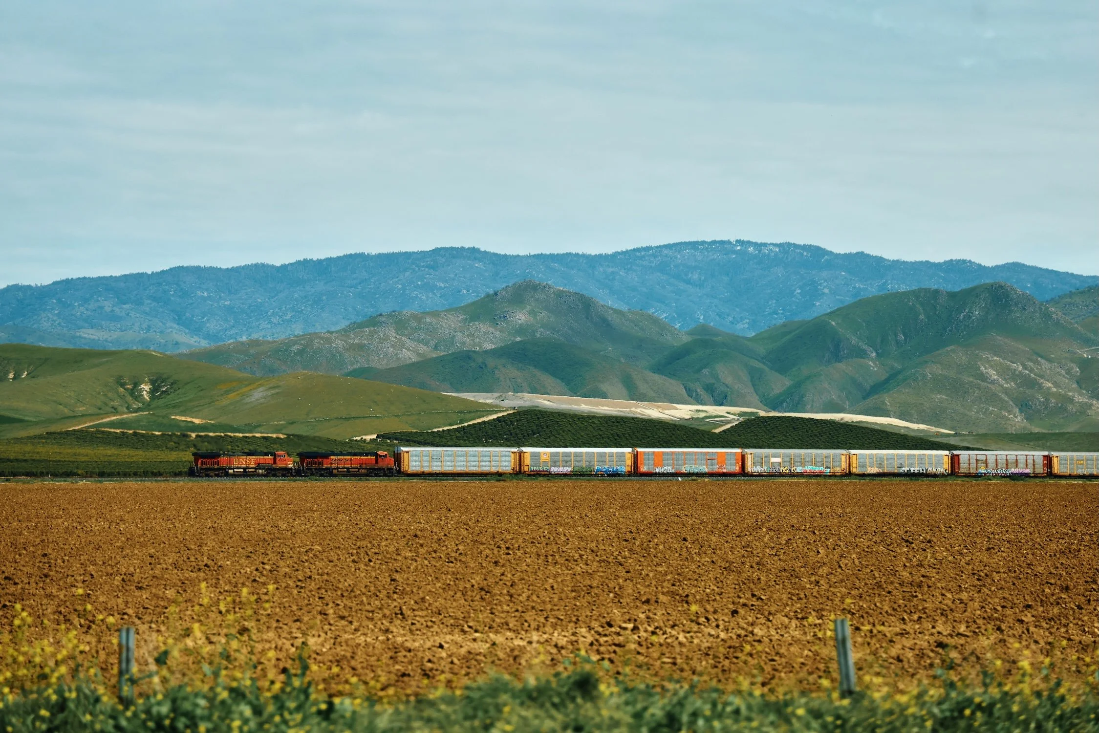A train with red, yellow, and orange cars running through a rural landscape with green hills and mountains in the background under a cloudy sky.