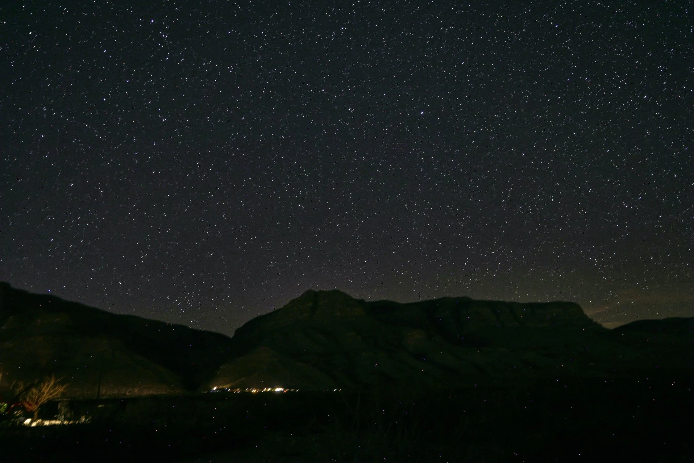 Night sky filled with stars over a hilly landscape with faint lights in the distance.
