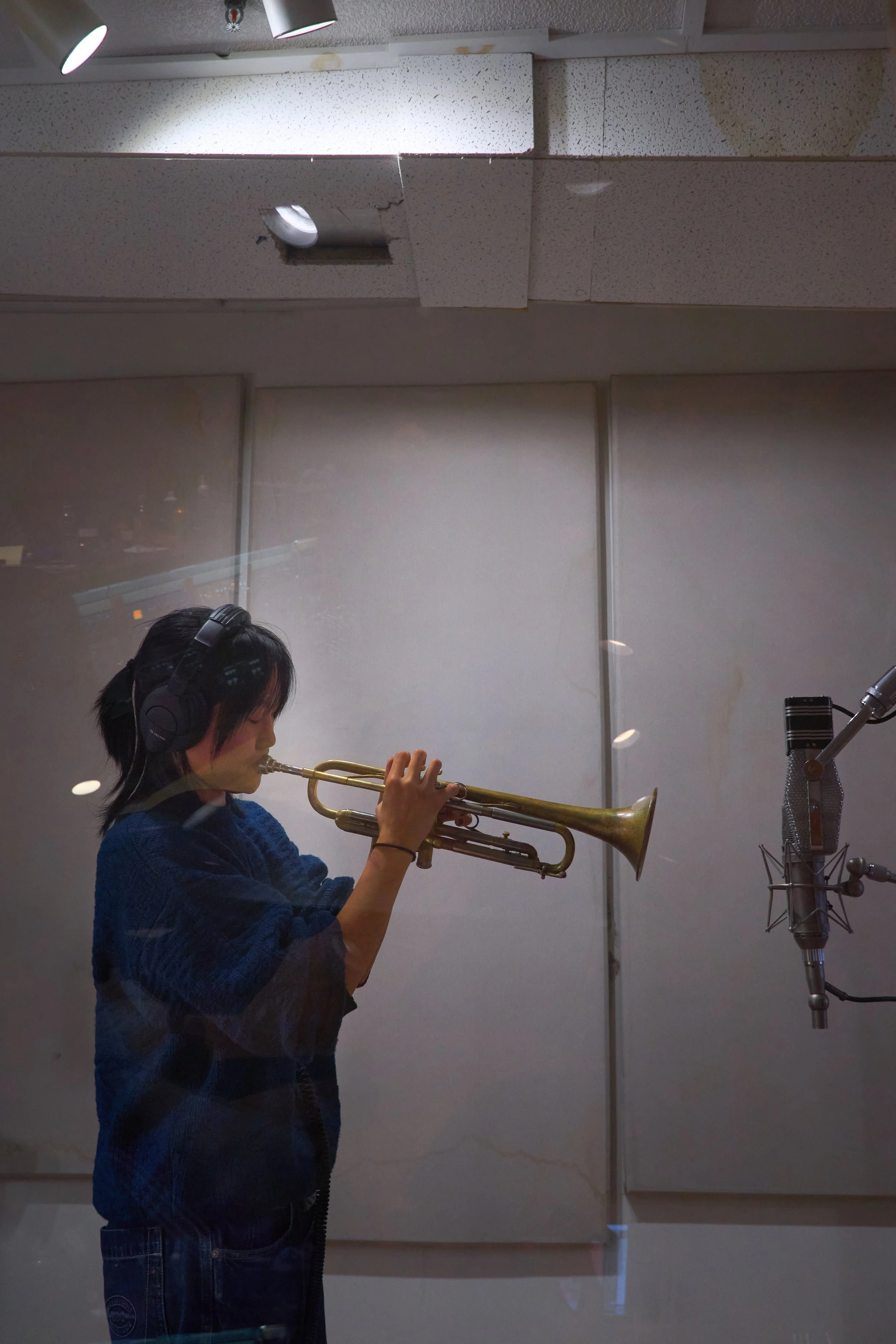Young person playing trumpet in a recording studio, wearing headphones, with a microphone set up in front of them.