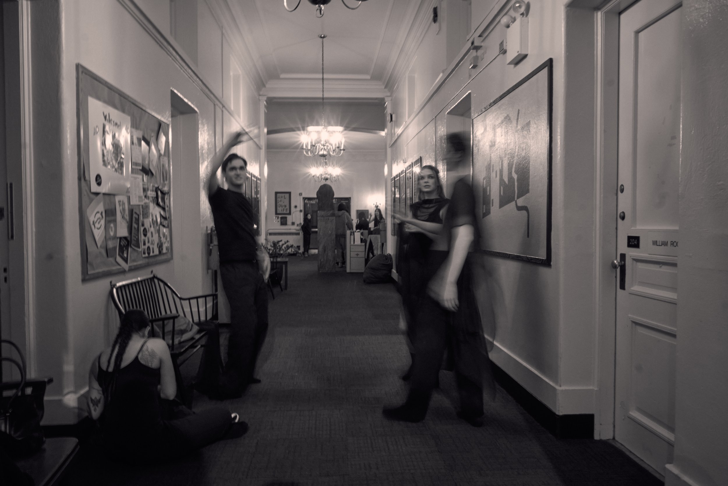 People standing and sitting in a hallway with lockers, bulletin board, and chandeliers, in a school or communal building.