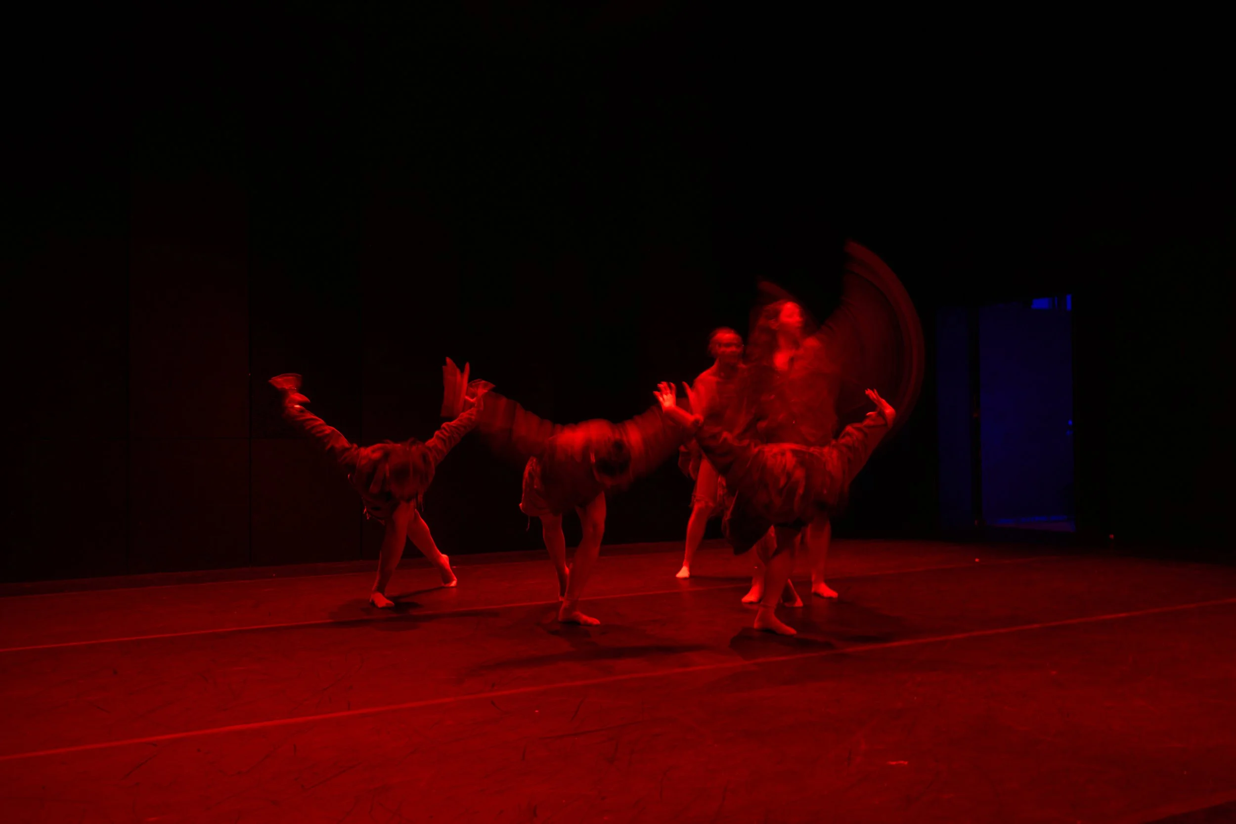 Group of dancers performing on stage illuminated with red light, some dancers are upside down or in dynamic poses.