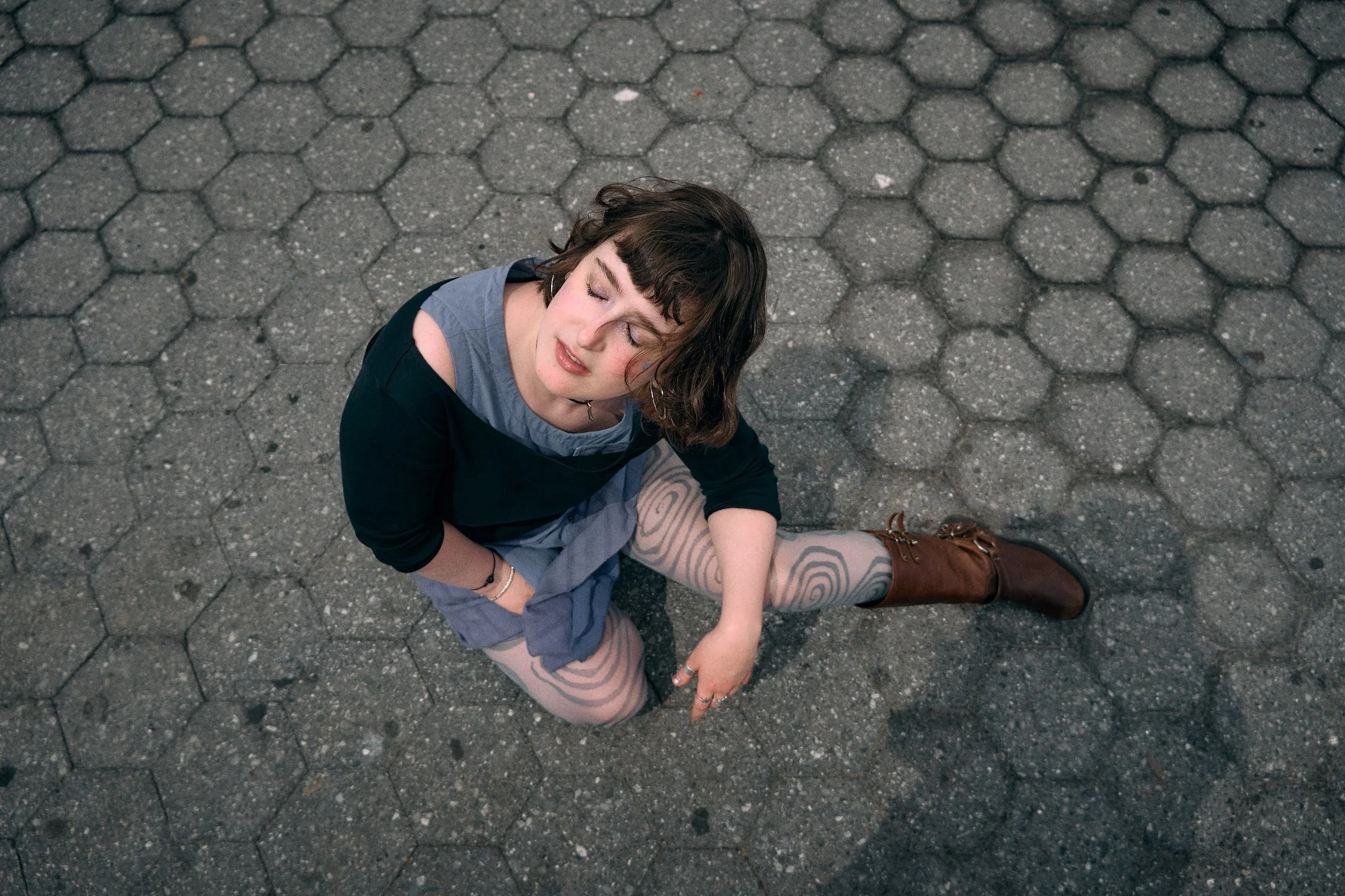 A young woman with curly brown hair and light skin sitting on a hexagonal patterned pavement, eyes closed, with a peaceful expression. She is wearing a black and gray top, patterned tights, and brown boots.