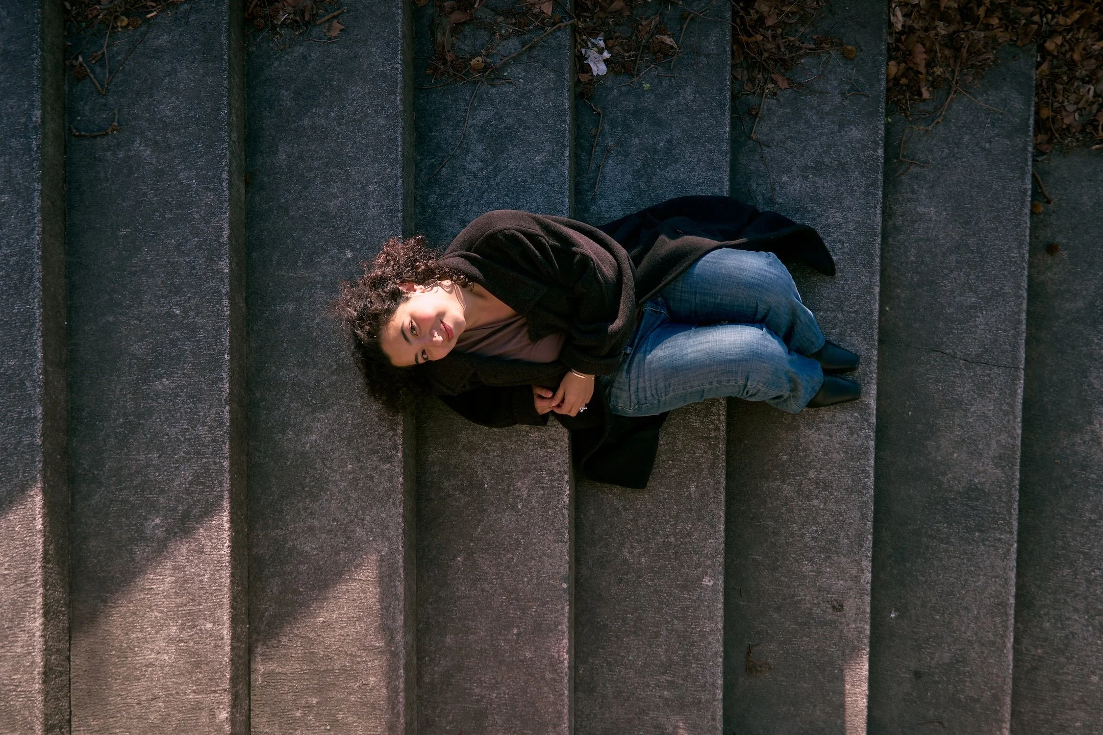A woman with curly hair lying on outdoor stone steps, wearing a black coat and jeans, looking up at the camera.