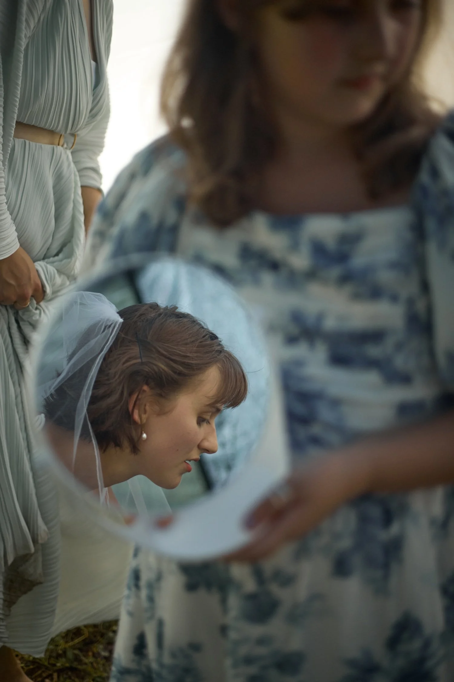 A woman in a wedding dress with a veil is clasping her hands and looking down, while a woman in a blue and white dress standing nearby is leaning over her, with her face partially obscured. A third woman in the background is wearing a white dress wit