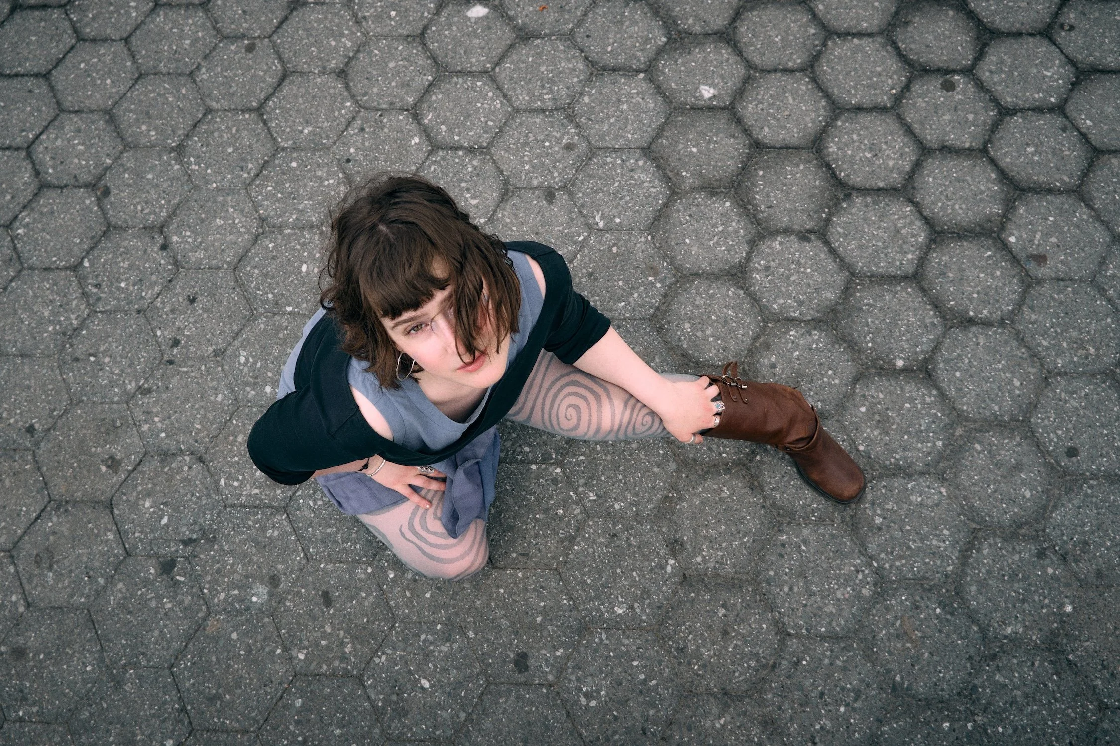 A young woman with short brown hair and bangs looking up at the camera with a slightly open mouth. She has a tattoo on her left leg. She is wearing a gray t-shirt, a navy blue jacket, hoop earrings, and brown boots. She is outdoors on a gray hexagona