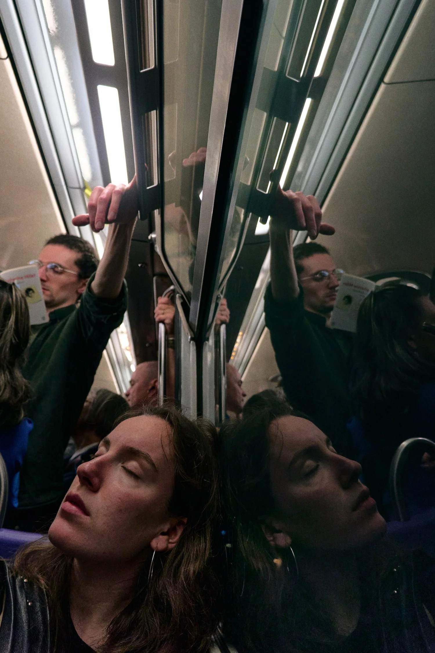 People sleeping with their heads resting against a reflective train door or window, some with eyes closed, in a crowded train or subway car.