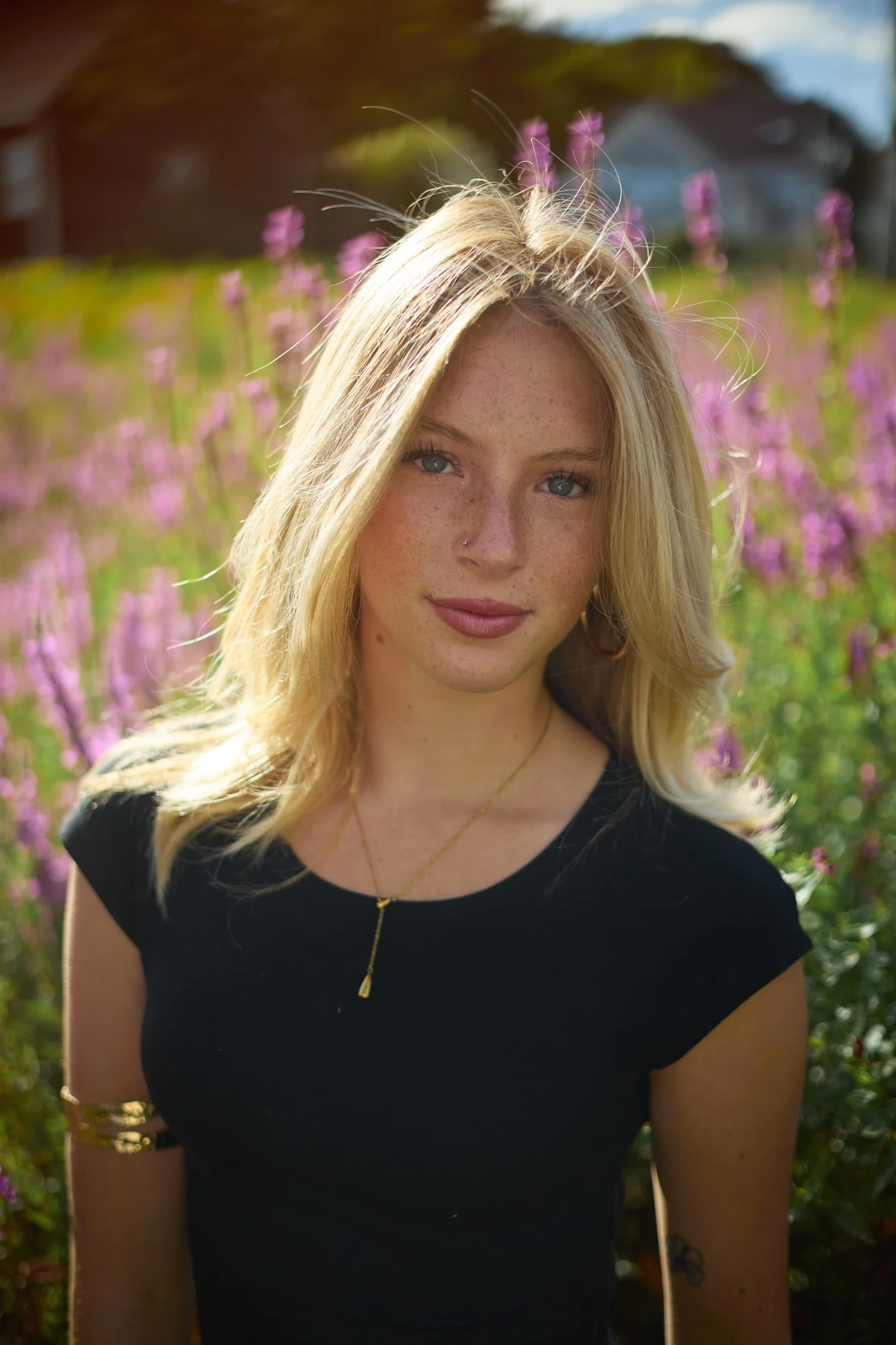 A young woman with blonde hair and freckles looking at the camera, standing outdoors in a field with pink flowers and a house in the background during daylight.