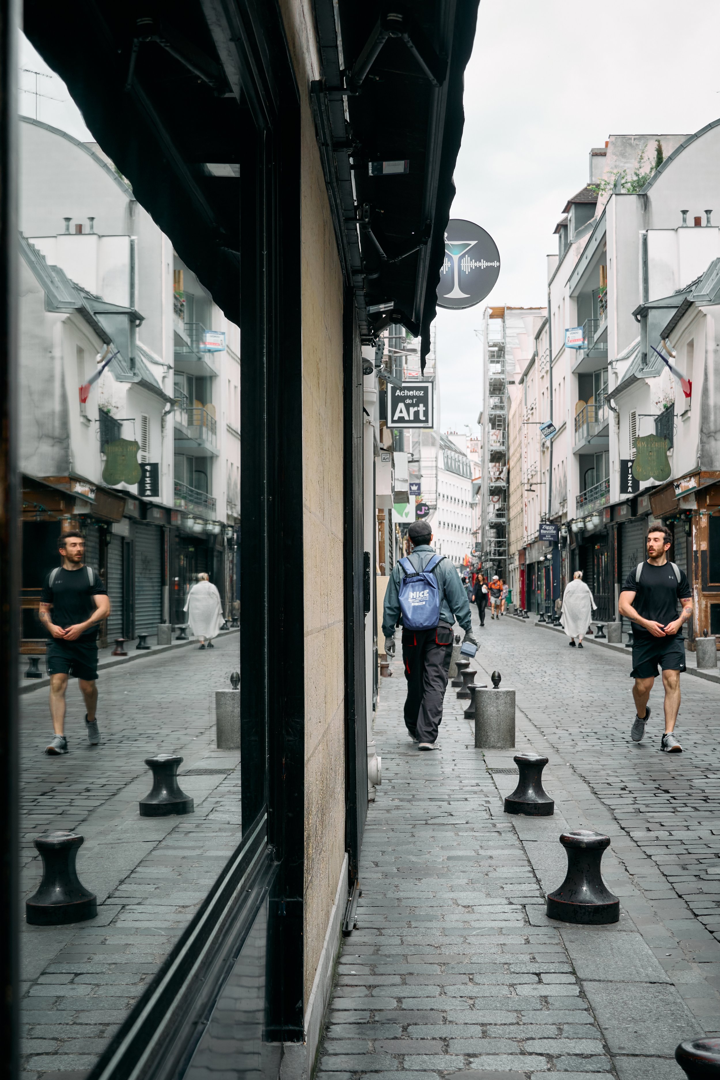 A city street with people walking on a cobblestone sidewalk, reflected in the glass window of a building, with various signs and buildings with balconies and graffiti in the background.