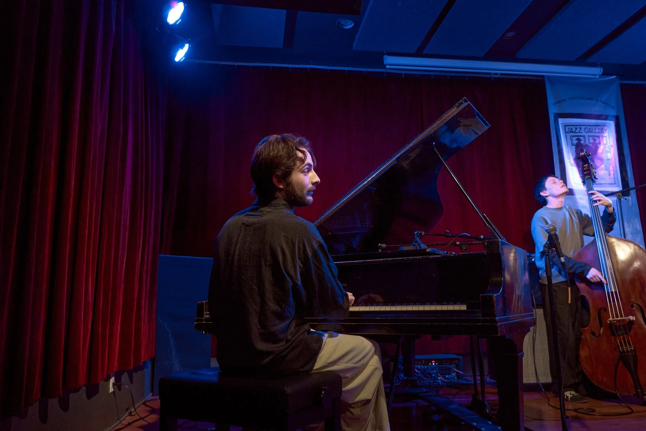 A man playing the piano on stage with red curtains in the background, under blue stage lighting, during a live jazz performance.