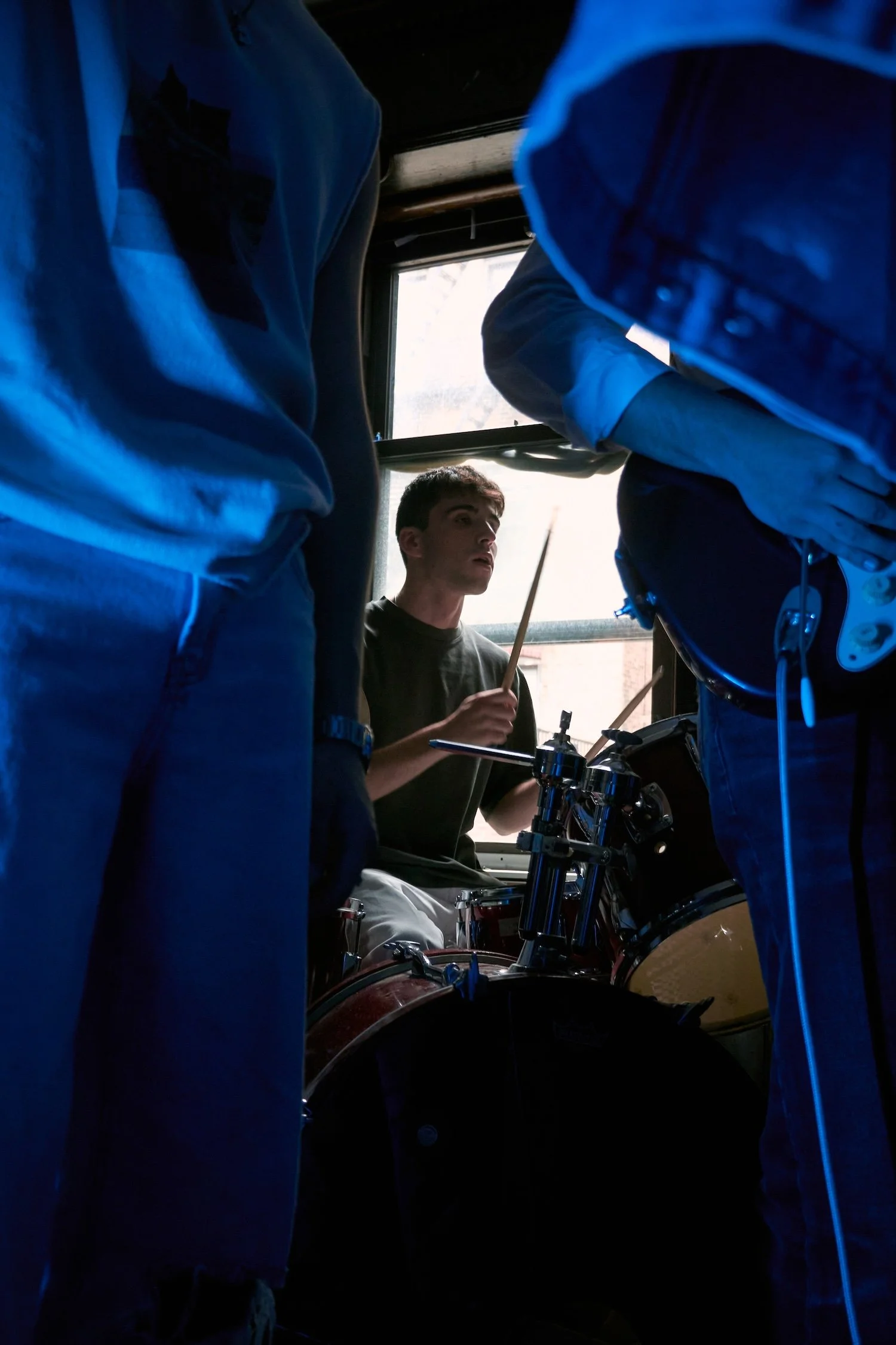 A young man playing drums in a dimly lit room with a window behind him, surrounded by band members.
