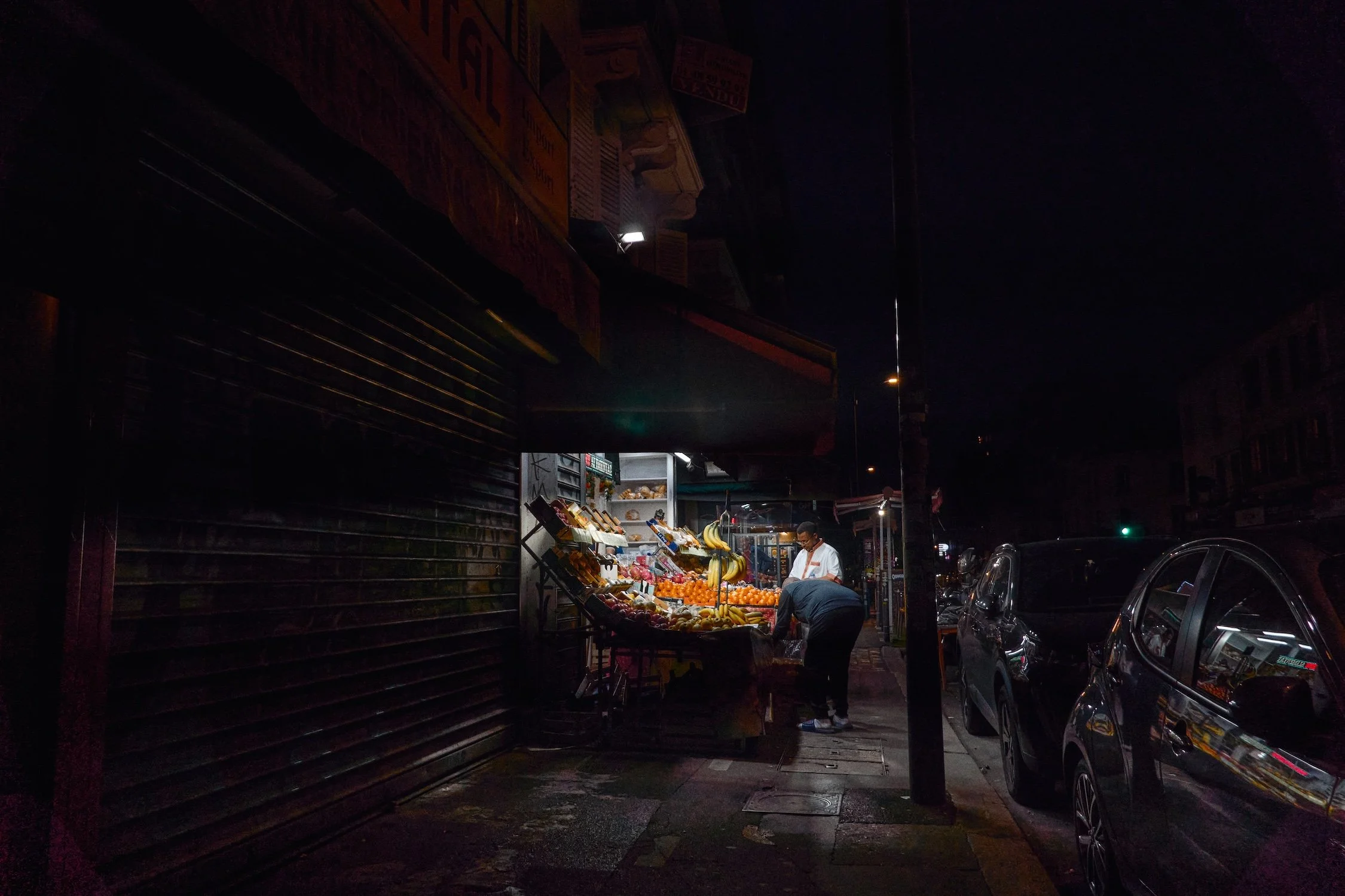 Nighttime street scene with a small fruit stand illuminated by a single light, with a person shopping and a closed shop behind a shutter, parked cars along the sidewalk, and dark buildings in the background.