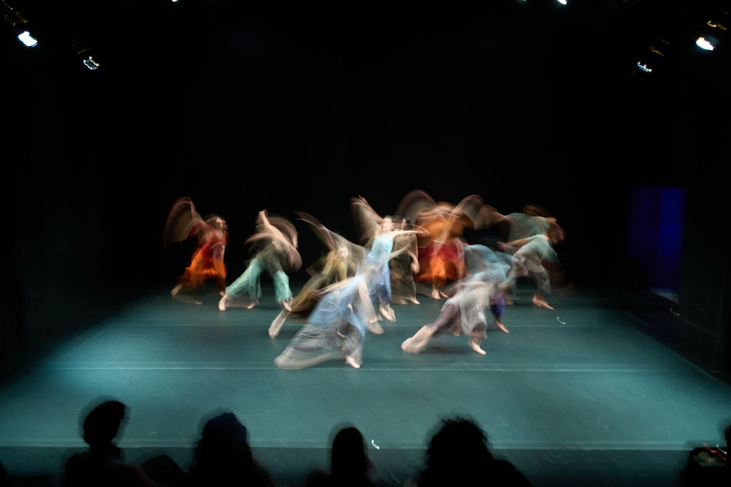 Blurred image of a group of dancers performing on stage in colorful costumes, with a dark background and audience members in the foreground.