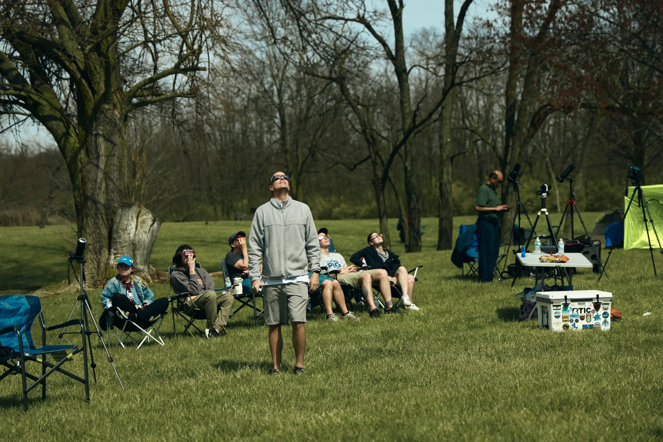 A man in a gray jacket and sunglasses looking up at the sky, standing outdoors on a grassy field during daytime with several people sitting on chairs around him, and multiple telescopes set up nearby. There are leafless trees in the background, indic