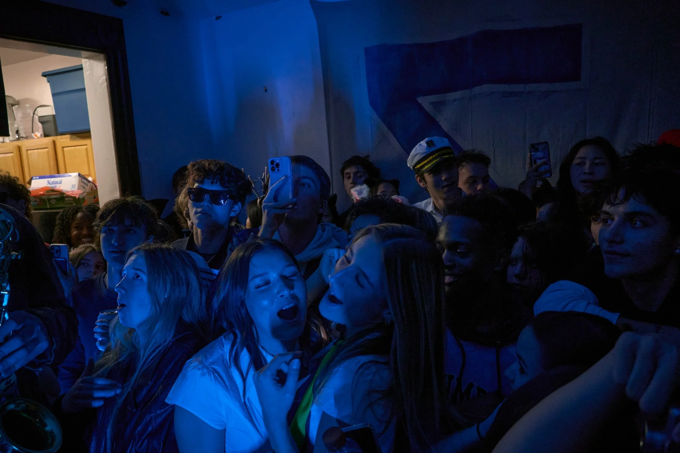 A group of young people at a dark indoor party, some holding drinks, with blue lighting, and a kitchen visible in the background.