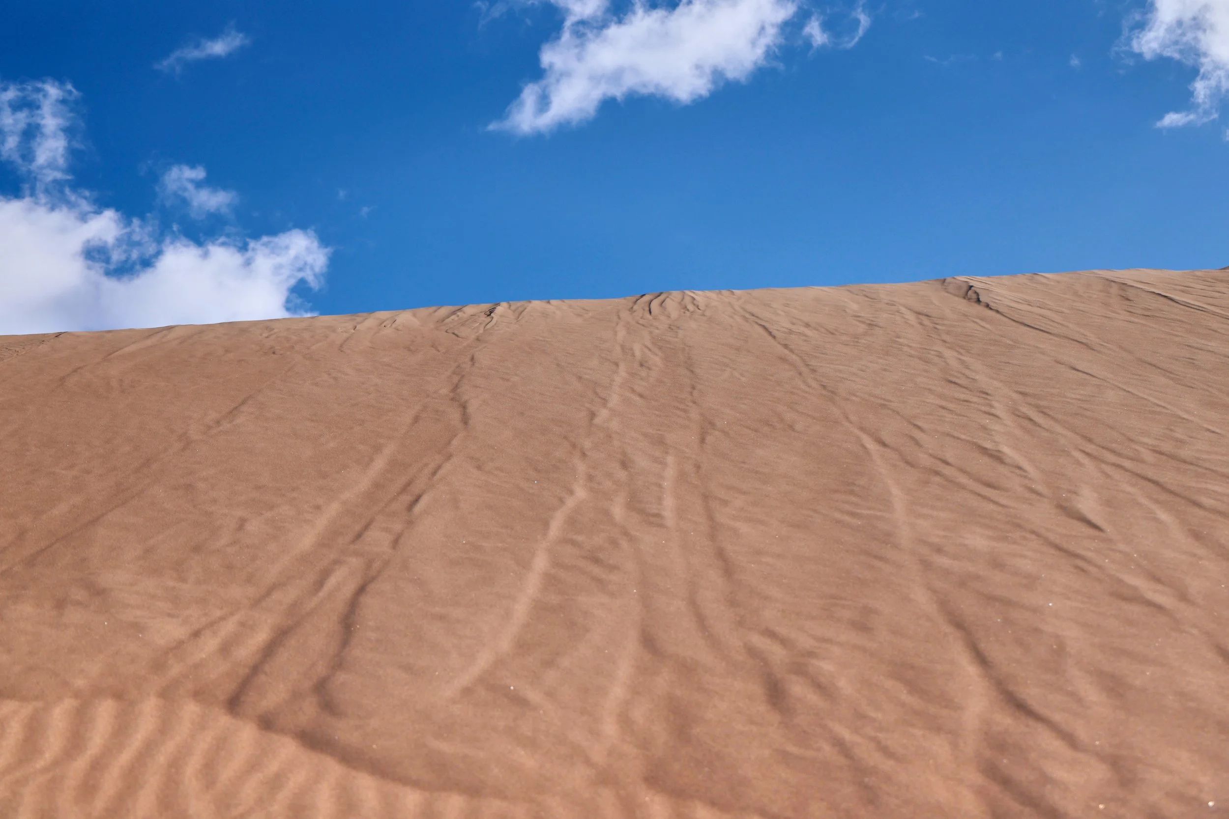 Close-up photo of a sandy surface with small ripples and textures, under a partially cloudy blue sky.