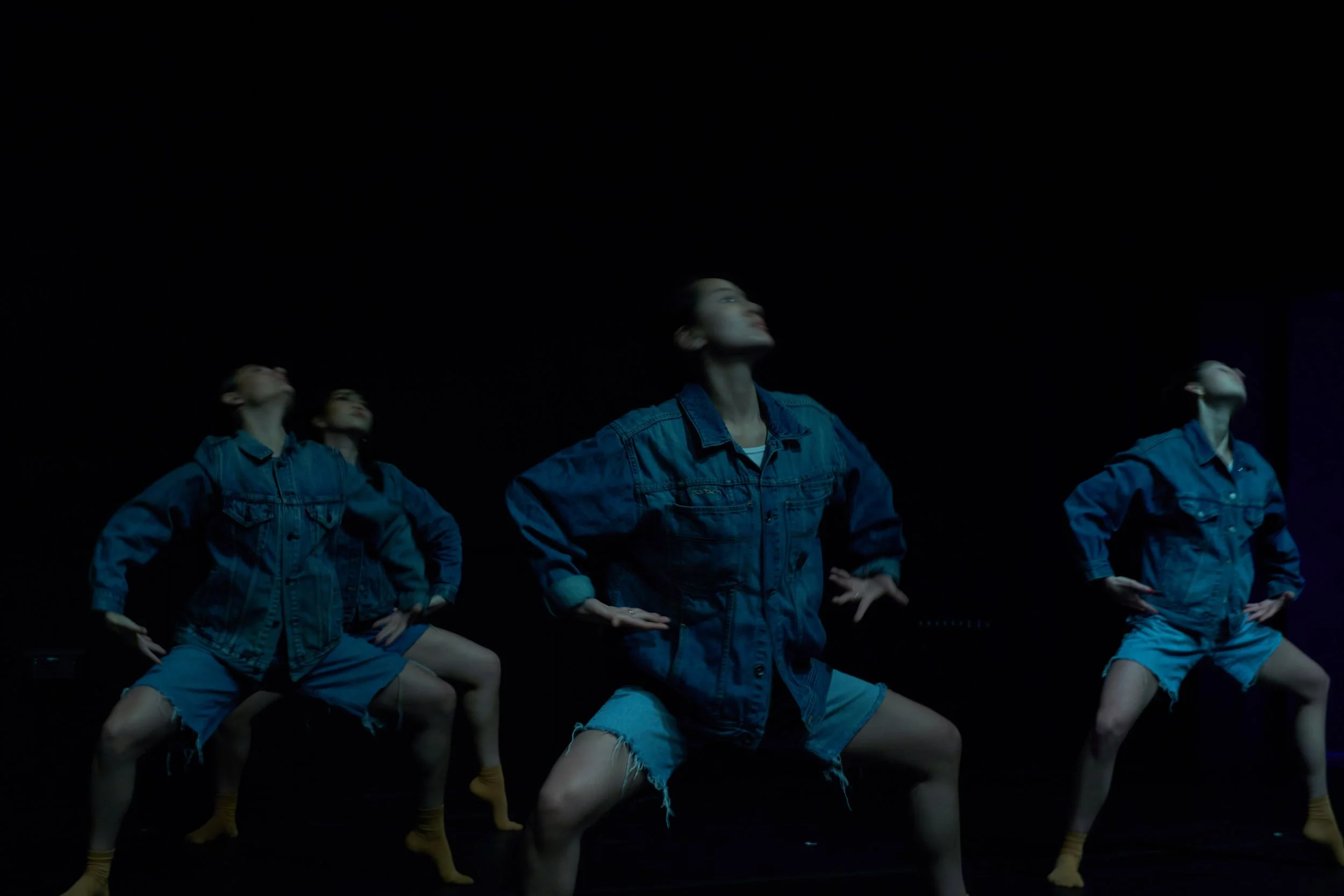 Five women in denim jackets and shorts posing with hands on hips, against a dark background.