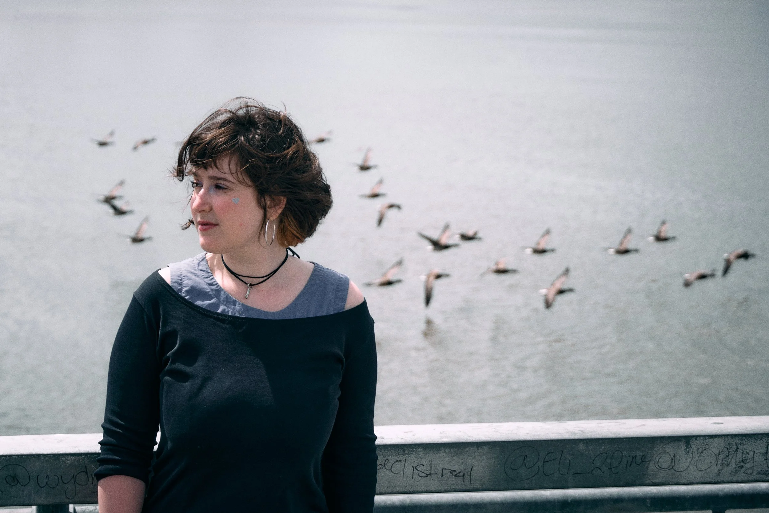 A young woman with short brown hair and hoop earrings standing outdoors by a body of water with seagulls flying in the background.