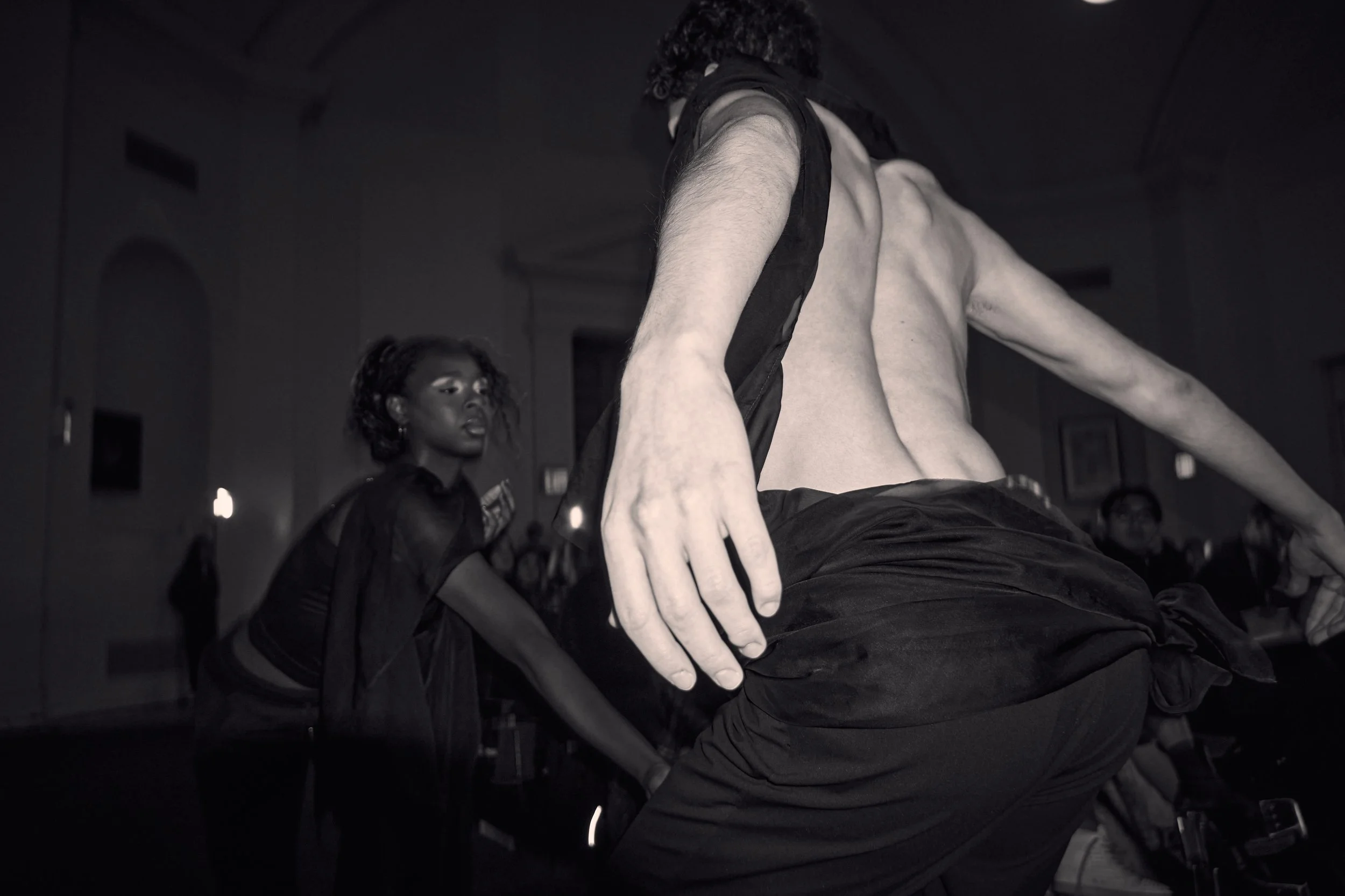 A Black woman and a man with curly hair in a dark room. The man is laying down and reaching out with his hand, while the woman stands behind him, looking at him.