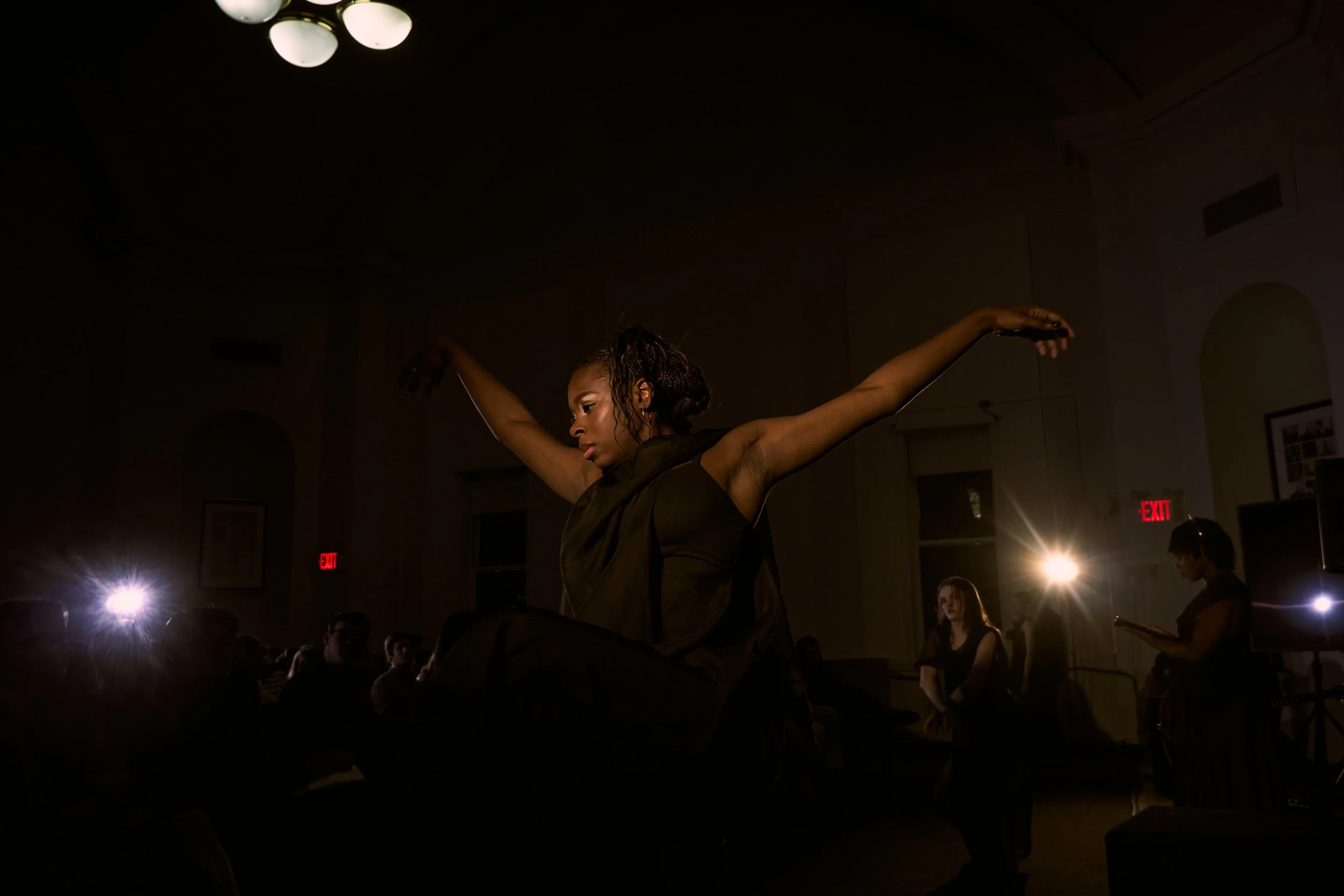 A woman with dark skin and curly hair performing a dance or pose with arms extended in a dimly lit room, audience watching, and bright lights and an exit sign visible.