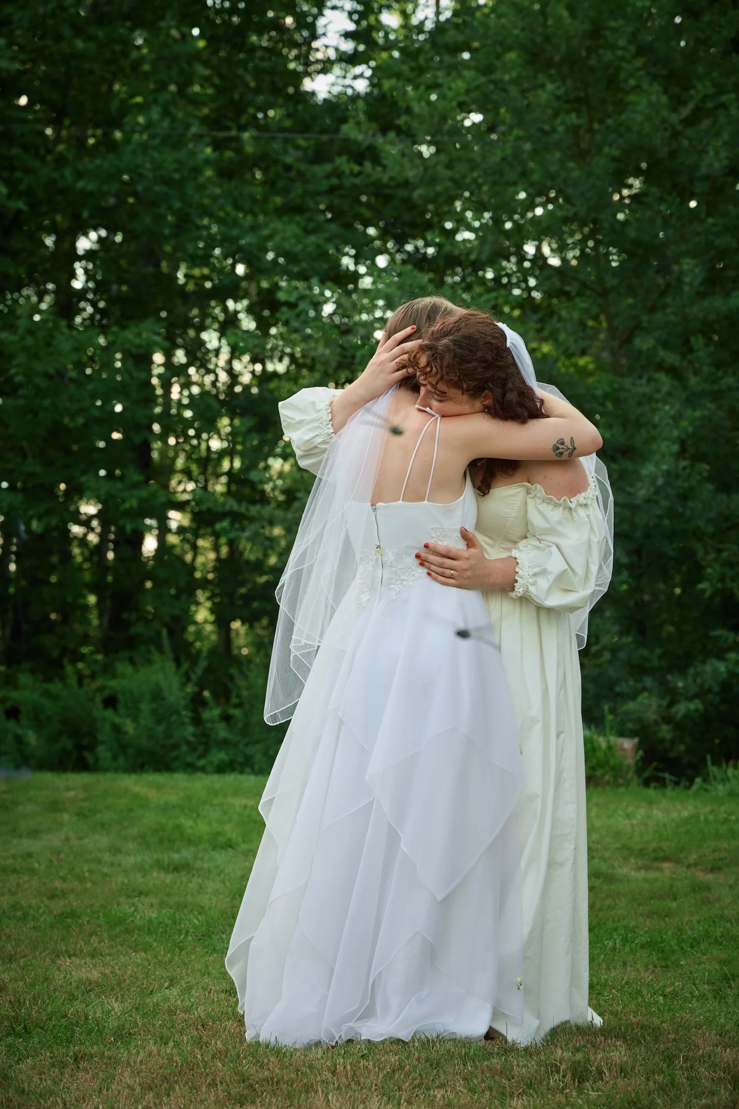 Two women in wedding dresses embracing outdoors in front of green trees, one with curly hair and a floral tattoo on her arm, the other with straight hair and wearing a veil.